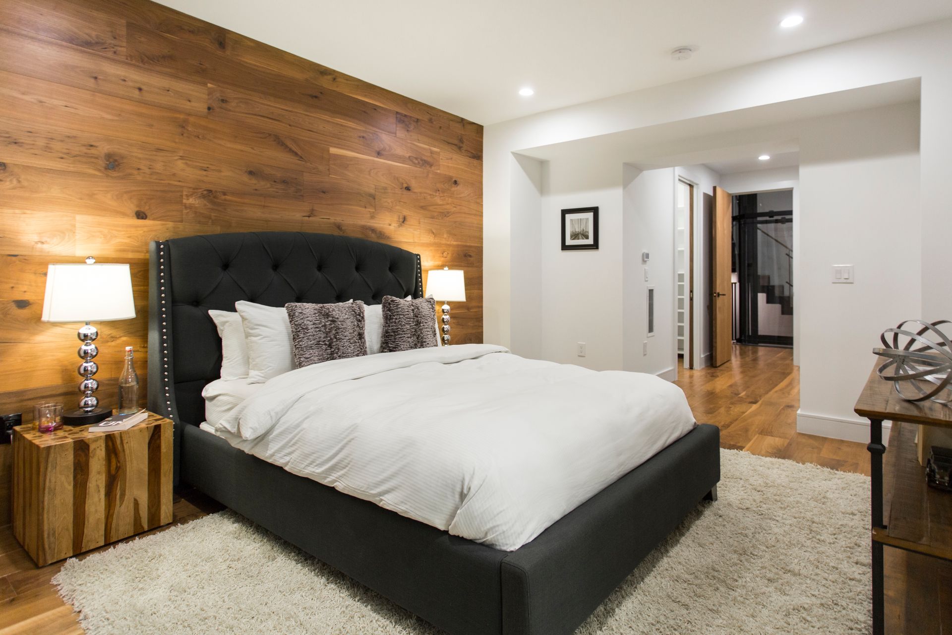 Bedroom with wood accent wall, gray bed, white bedding, fluffy rug, and lamps.