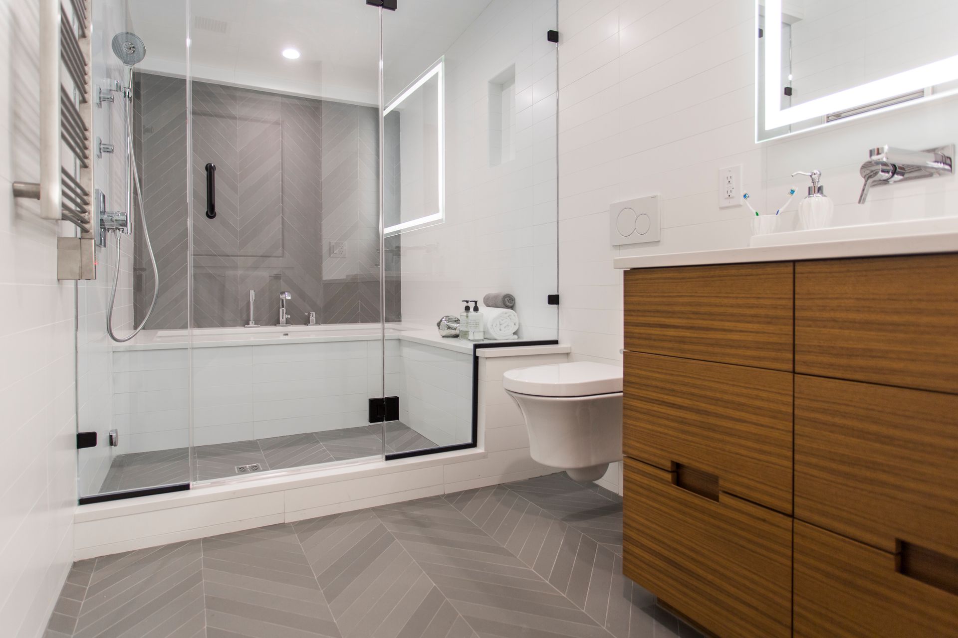 Modern bathroom with glass shower, wooden vanity, white toilet, and gray herringbone floor.