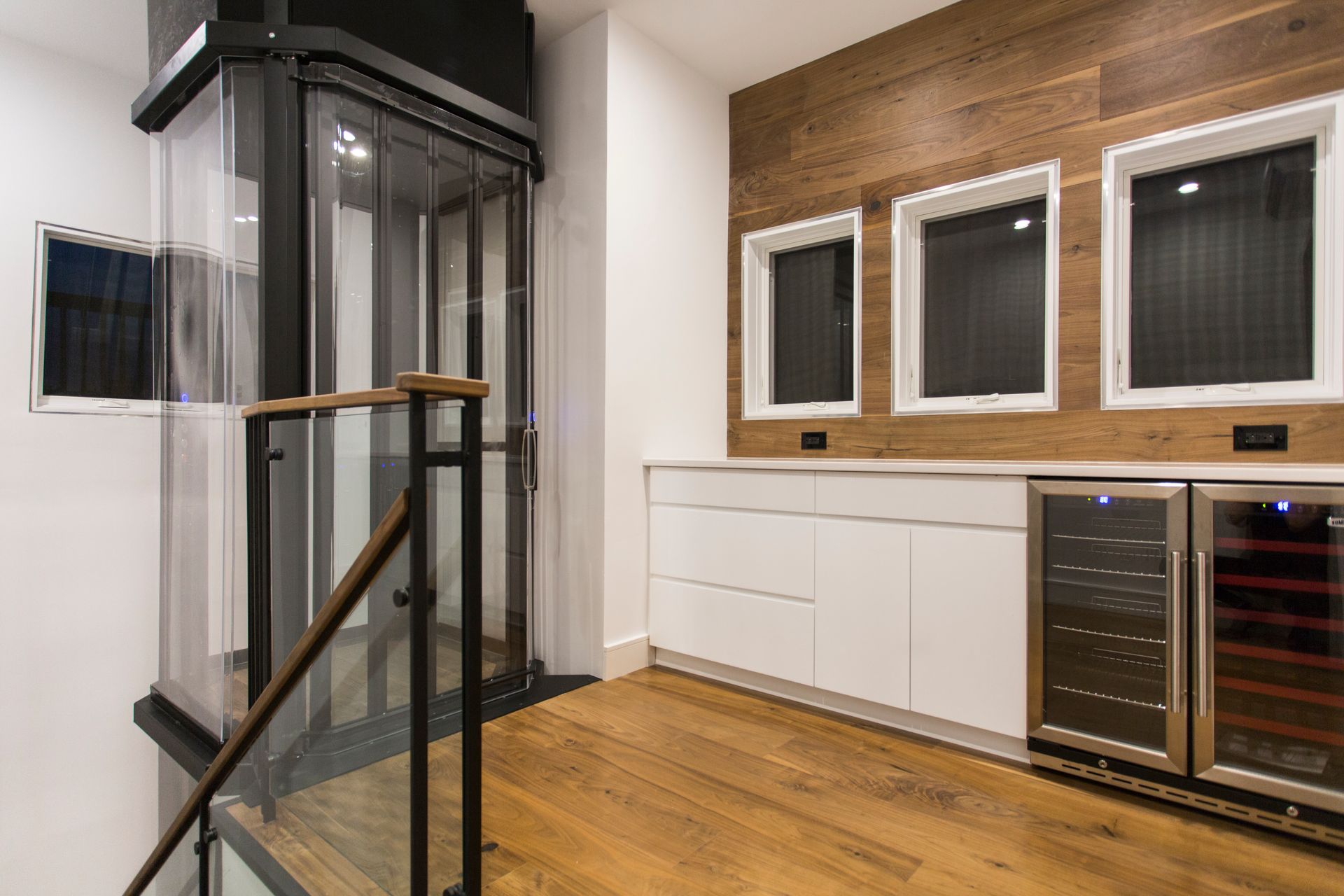 A modern home interior with a glass elevator, wood paneling, white cabinets, and wine fridges.