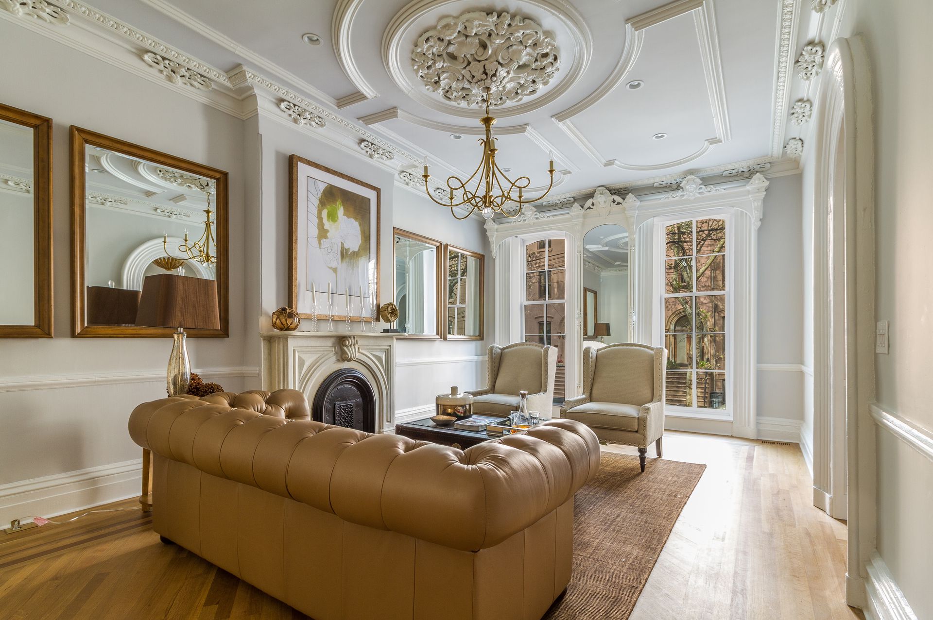 Living room with ornate ceiling, fireplace, tan sofa, chairs, and mirrors.