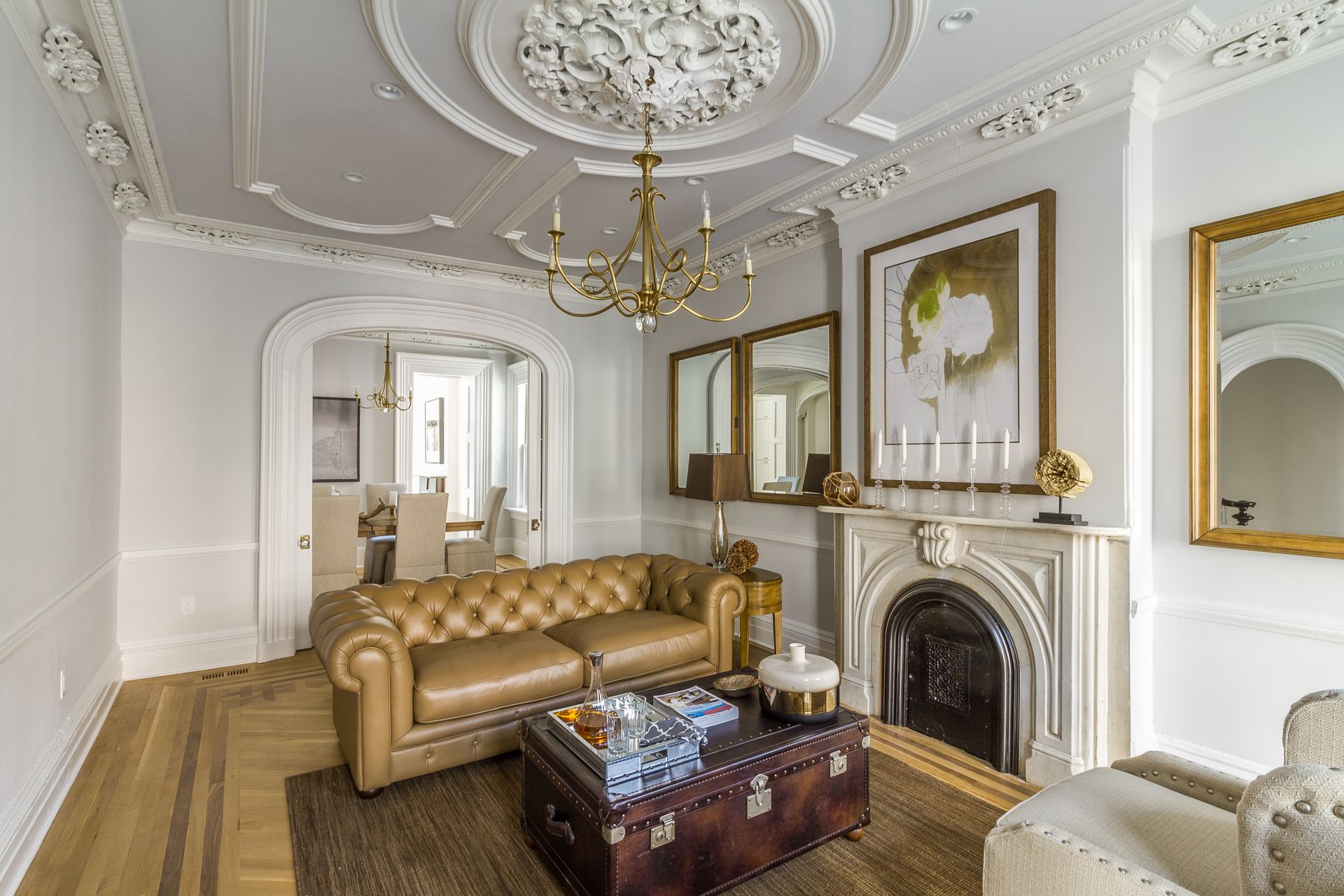Living room with ornate ceiling, fireplace, leather sofa, wood floors, mirrors, and a decorative trunk.