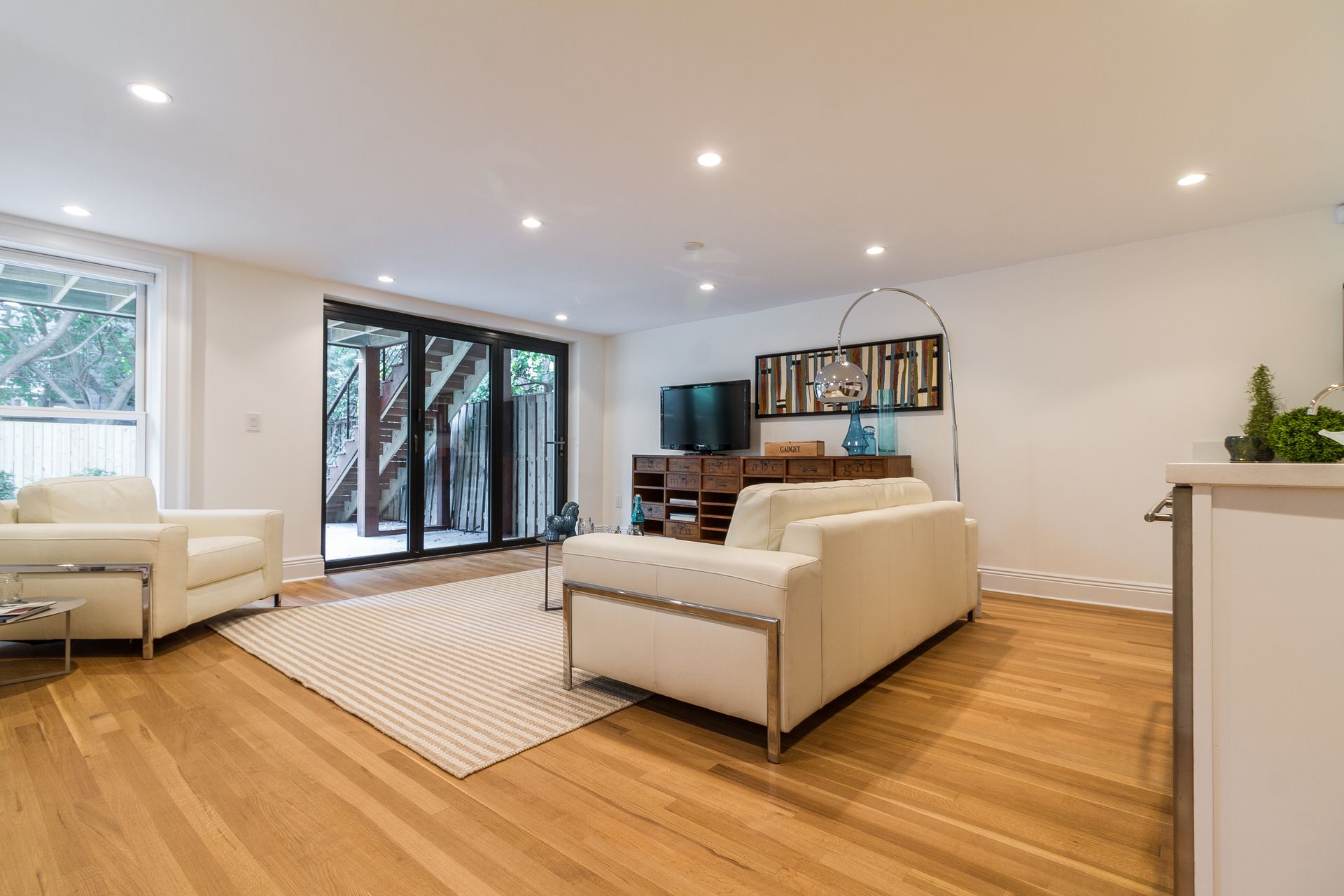 Modern living room with white furniture, wooden floors, and large glass doors leading to a backyard.