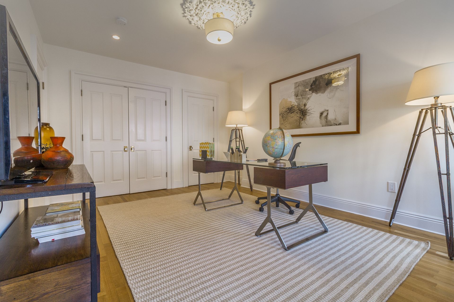 Home office with desk, rug, globe, and artwork. Features white walls, doors, and a decorative ceiling light.