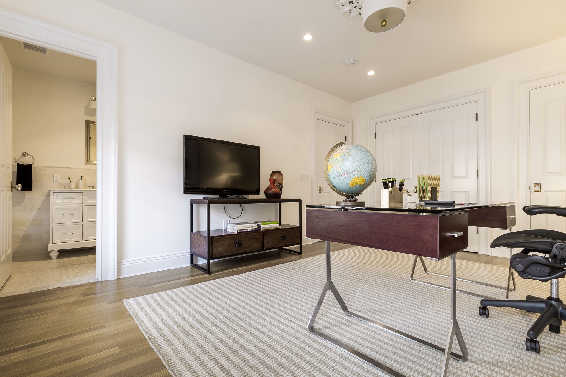 Home office with desk, TV, globe, and two doors. Light walls, wood floor, and cream rug.