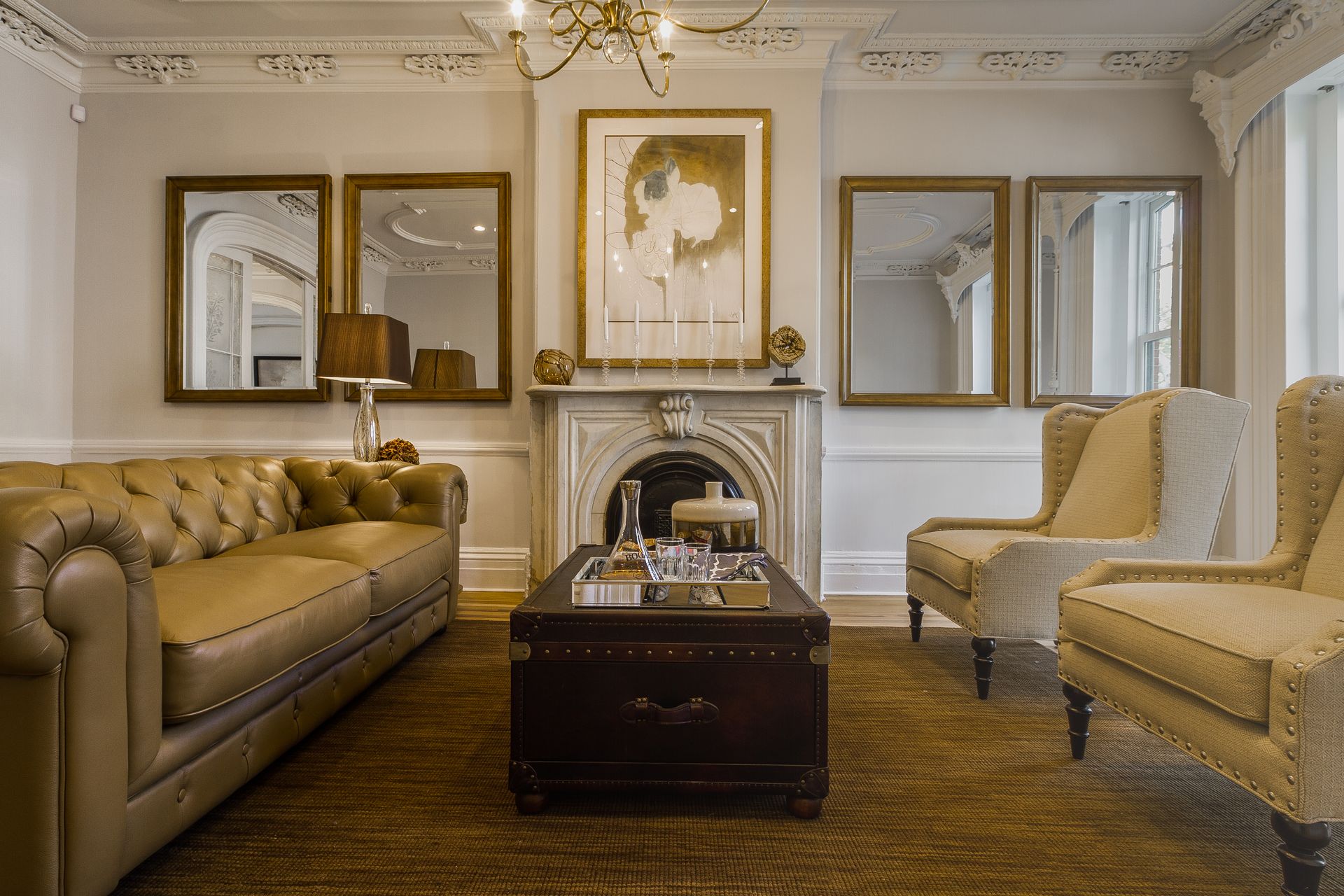 Elegant living room with leather sofa, two armchairs, decorative fireplace, mirrors, and coffee table on a woven rug.