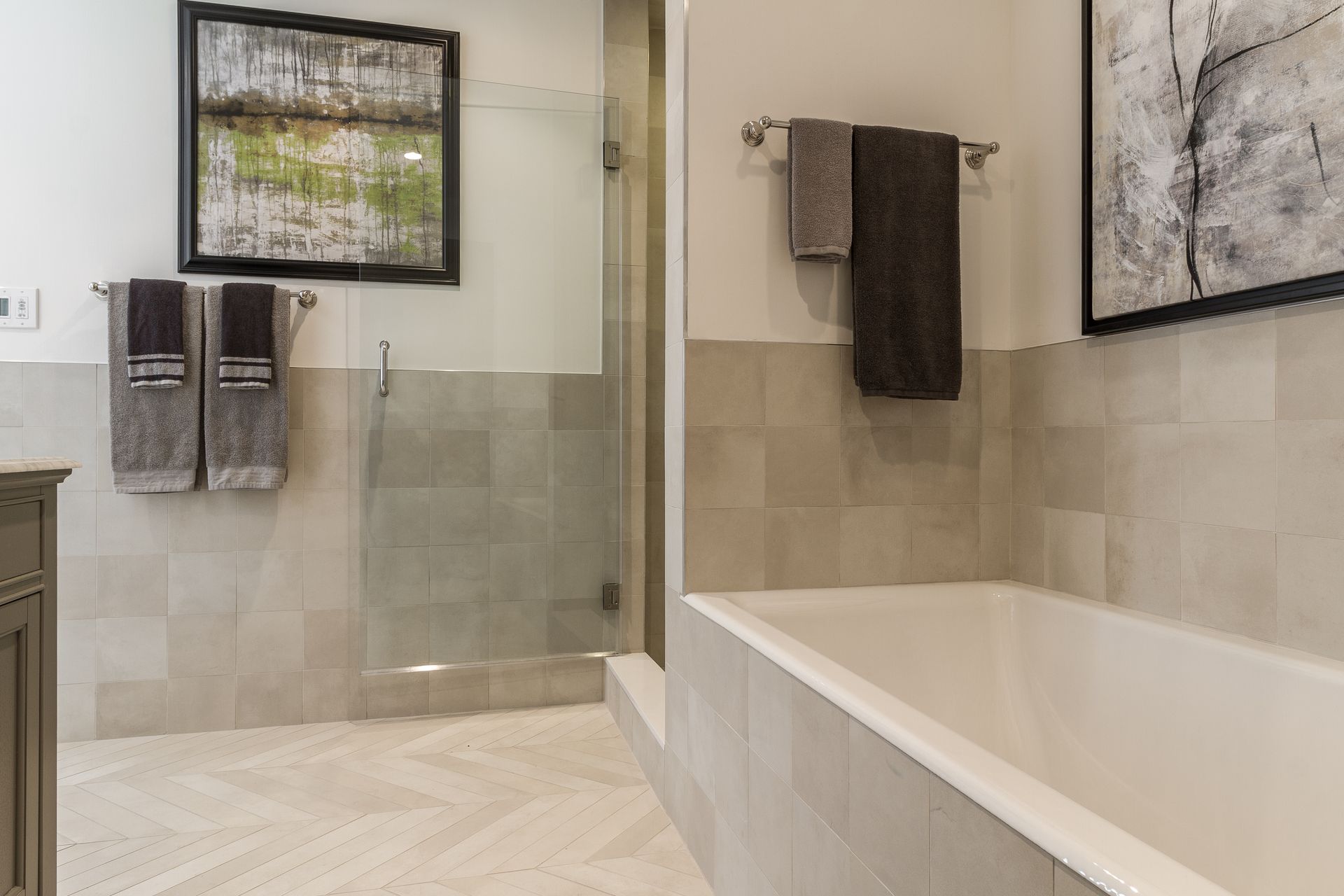 Modern bathroom with a bathtub, shower, neutral tile, and framed art. Two sets of towels hang on the walls.