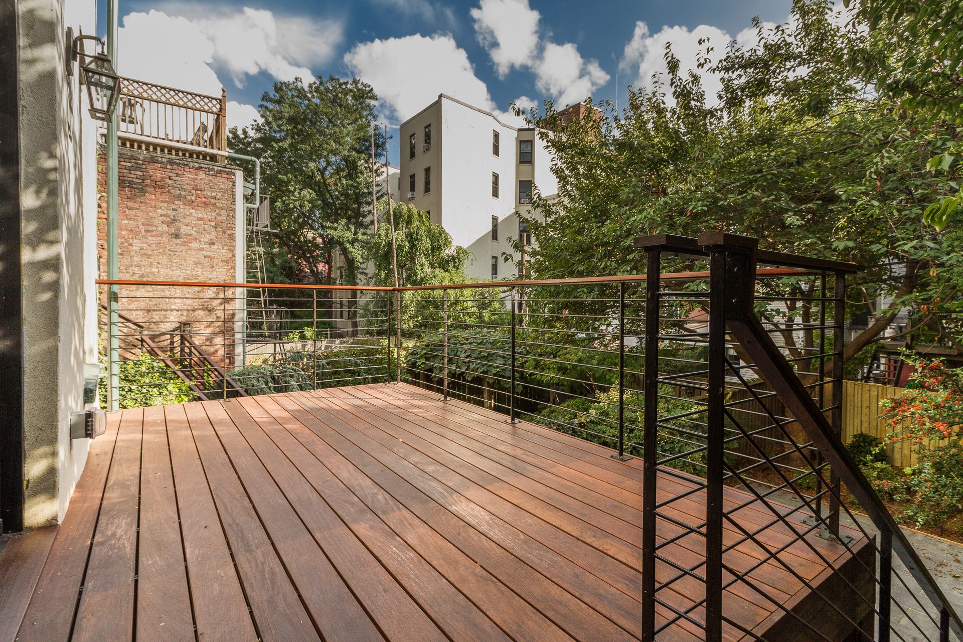 Wooden deck with metal railings overlooking a backyard with trees and a brick wall.