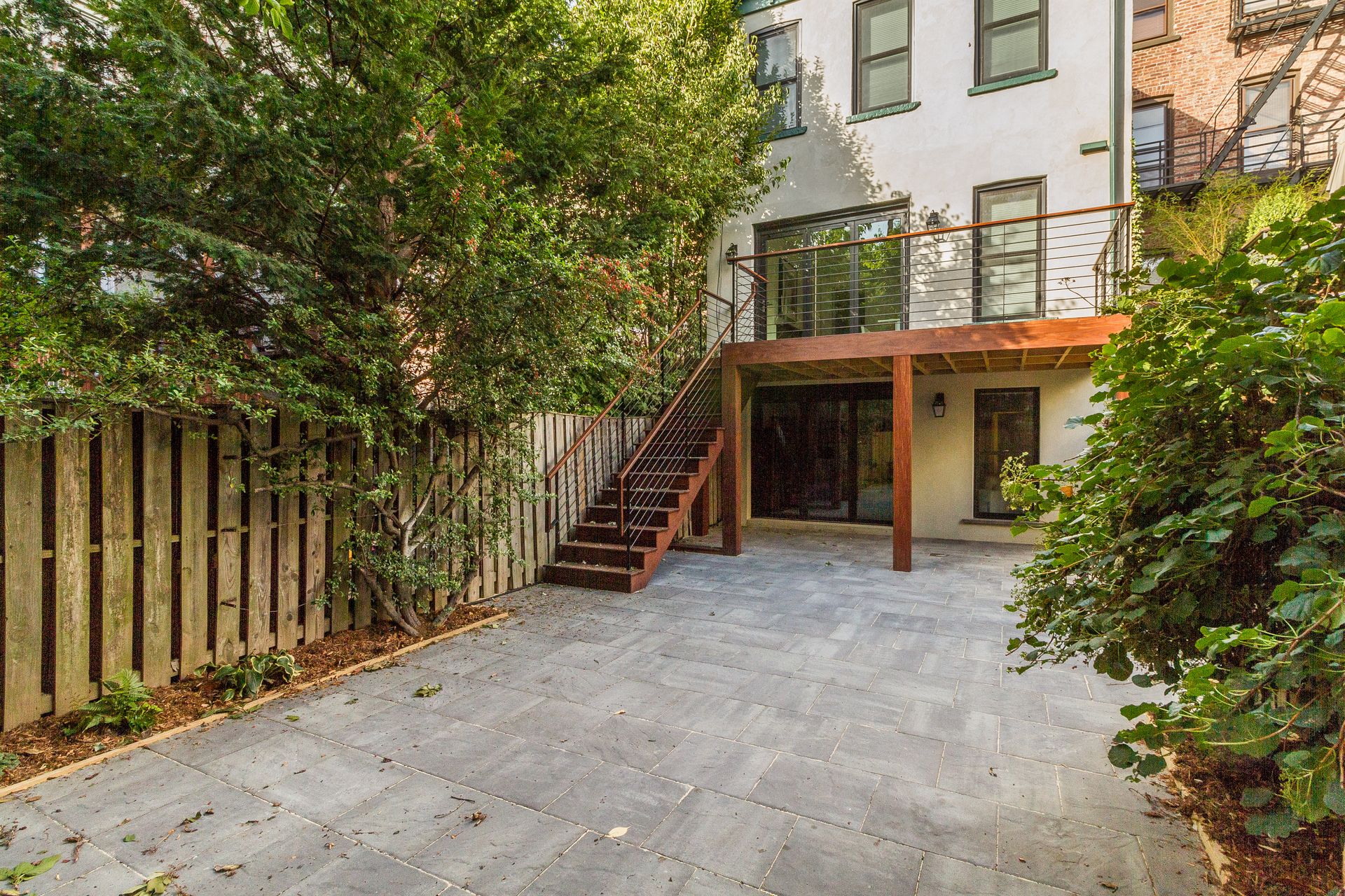 Backyard with gray pavers, wooden stairs, and a white building with a deck.