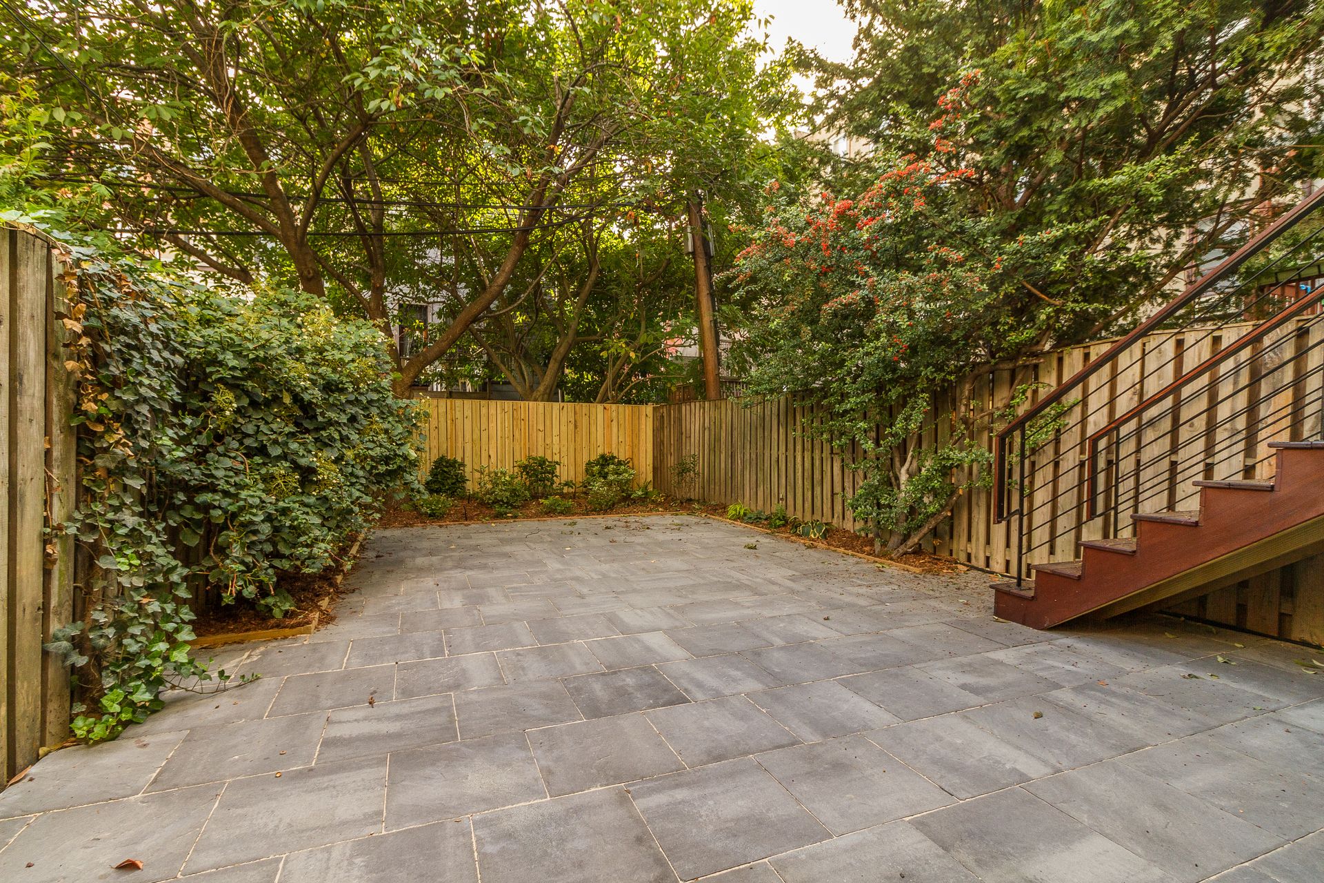 A paved backyard with a wooden fence, green foliage, and a staircase.