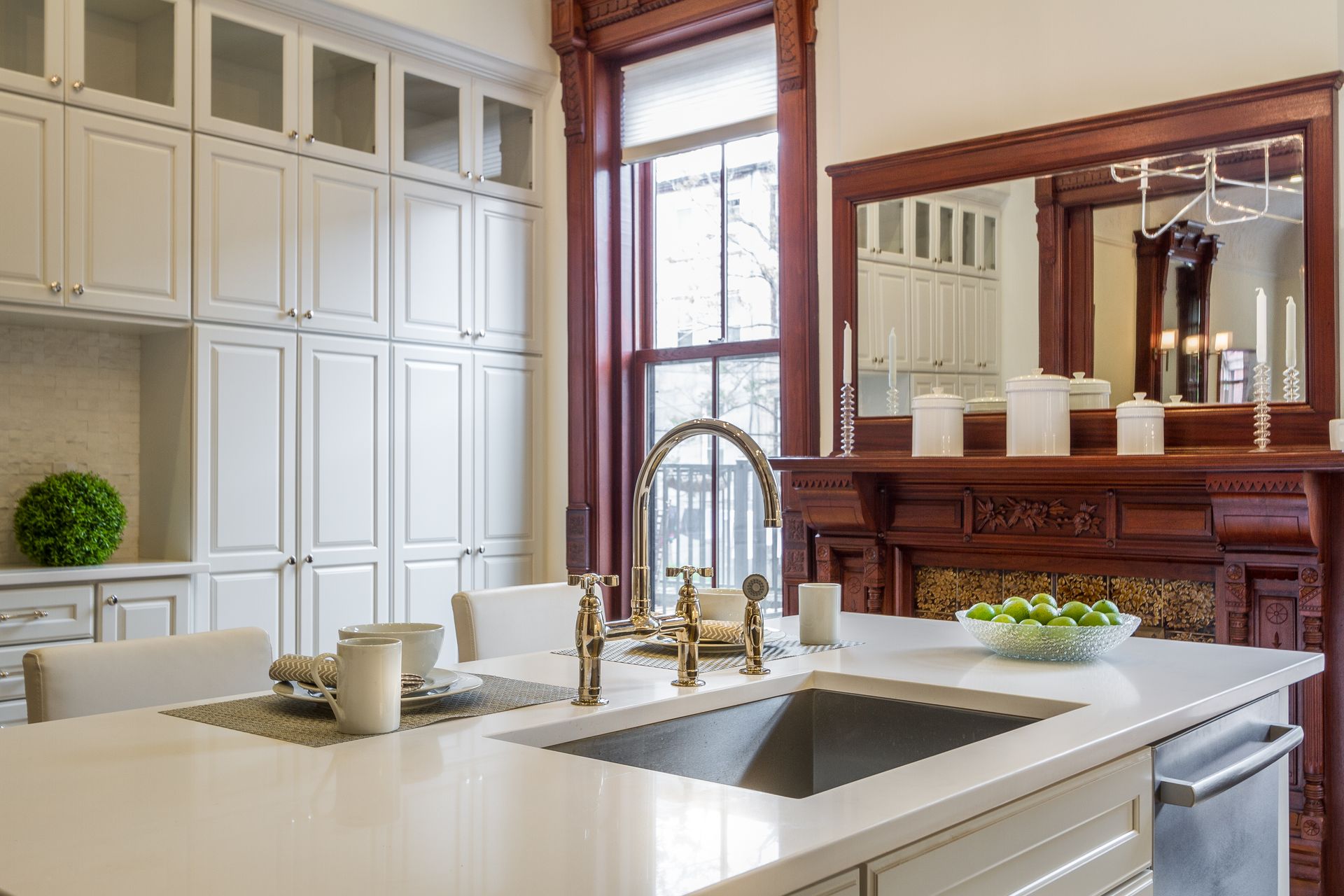 Kitchen with white cabinets, island sink, and ornate dark wood trim around a window and fireplace.
