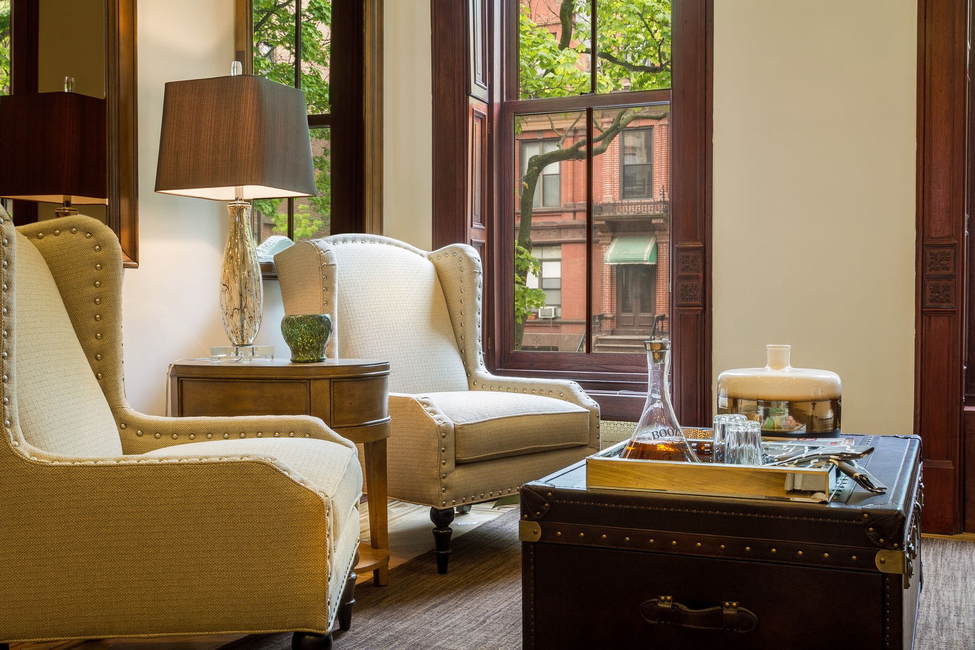 Cozy living room with two armchairs, lamp on side table, and a trunk coffee table. View of brick building through window.