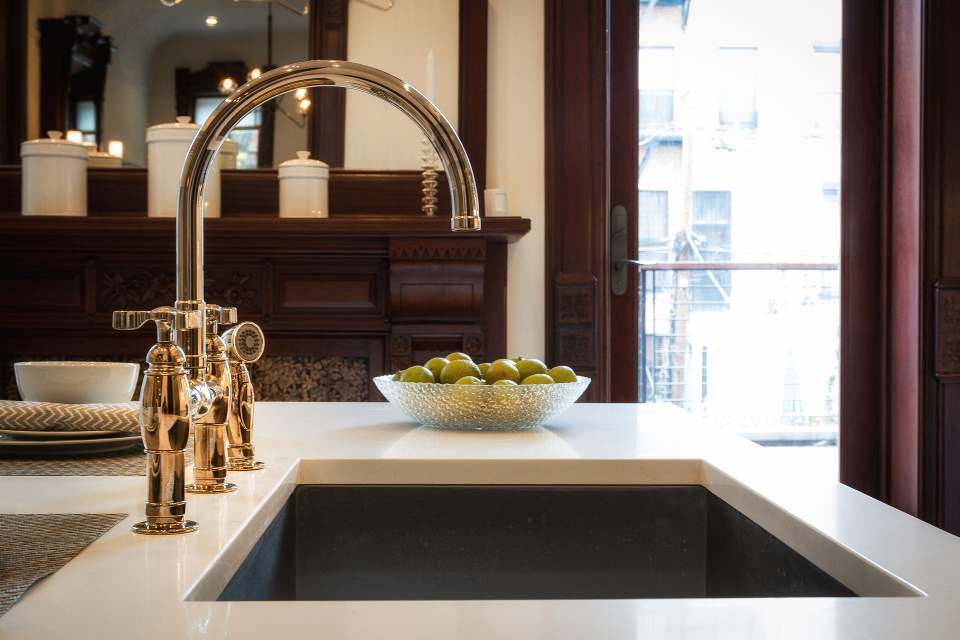 Kitchen sink with gold faucet, white countertop, and bowl of limes. Dark wooden trim surrounds window.