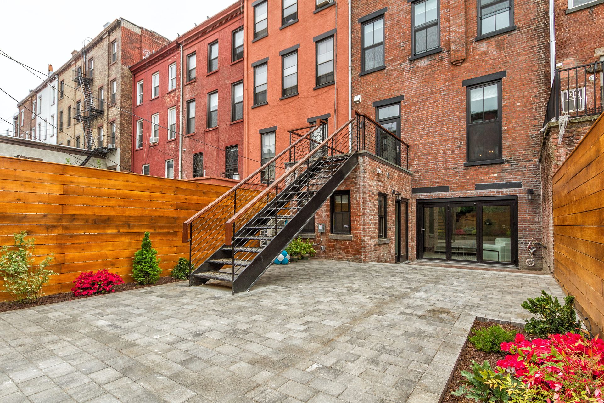 Backyard with brick buildings, metal staircase, wooden fence, and paved patio.