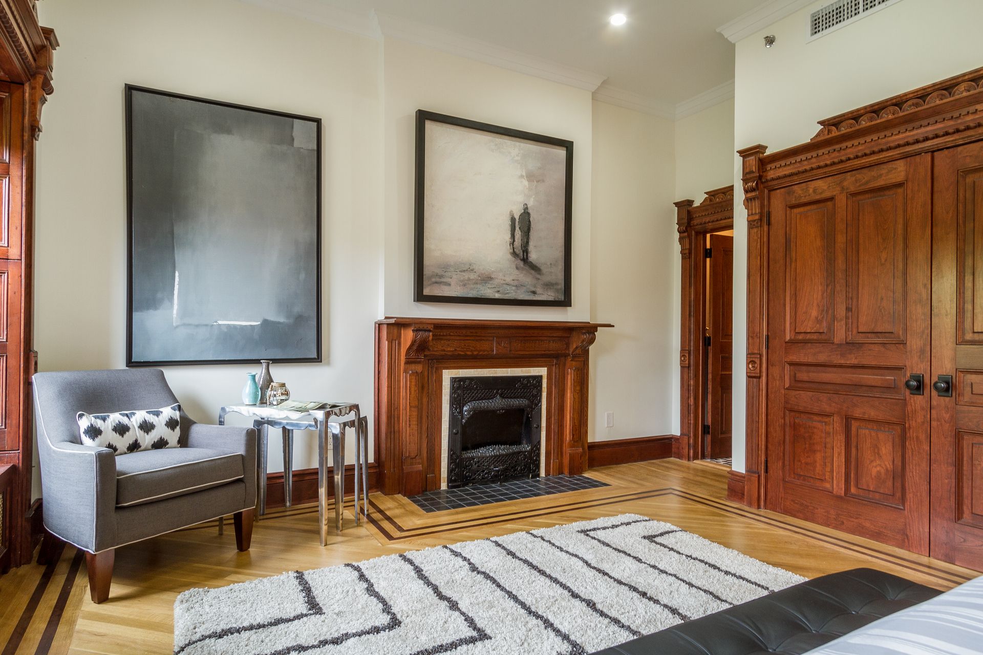 Bedroom with gray chair, fireplace, artwork, wood trim, and a patterned rug.