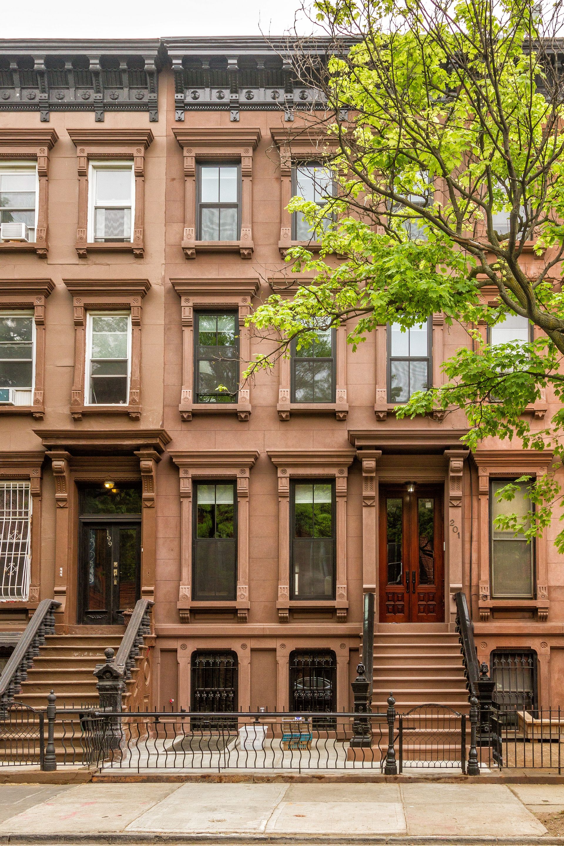 Brownstone buildings with steps, windows, and decorative ironwork. A tree with green leaves is on the right.