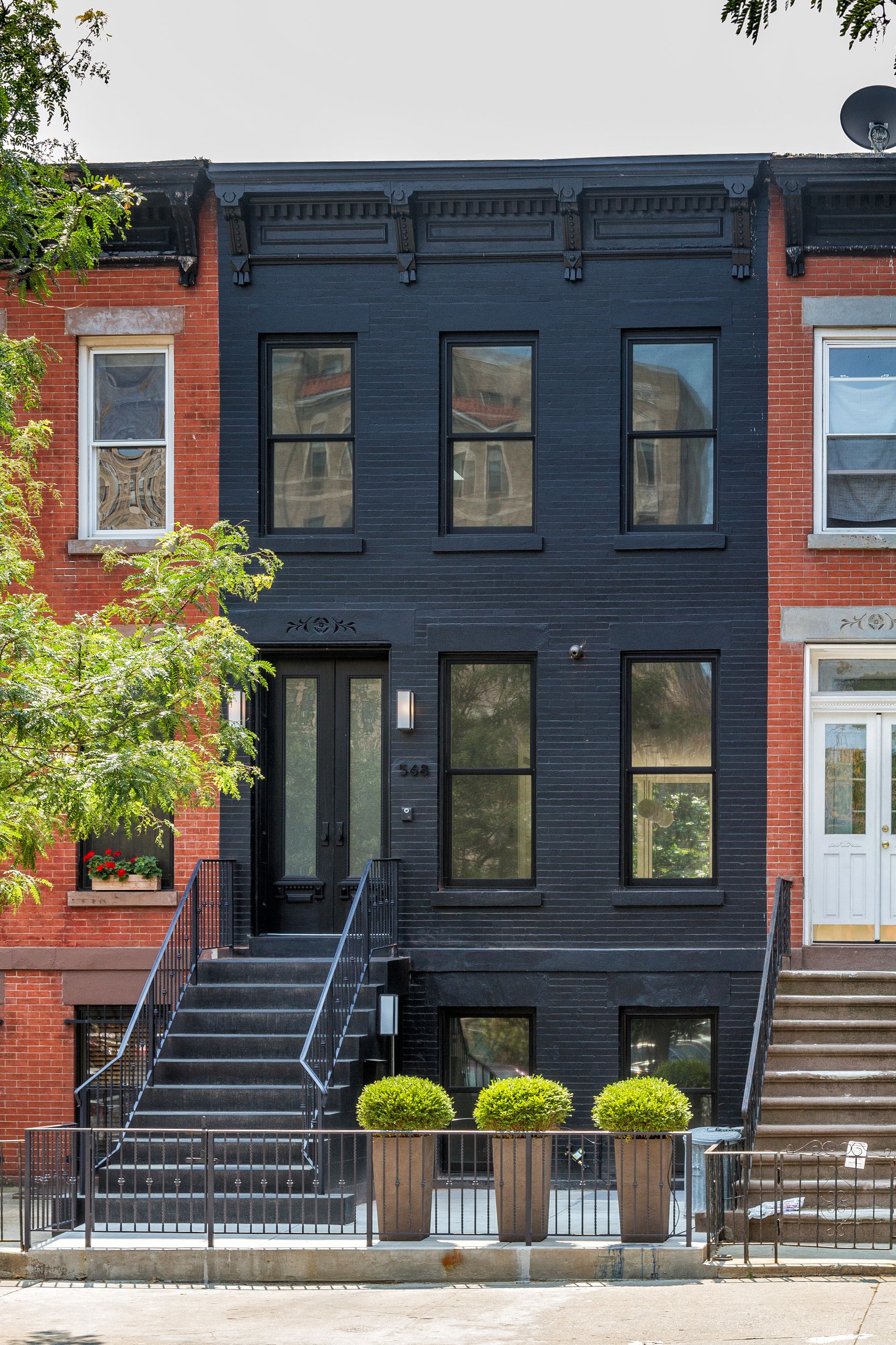 Black townhouse with black framed windows, front steps, and potted plants in front of a metal fence.