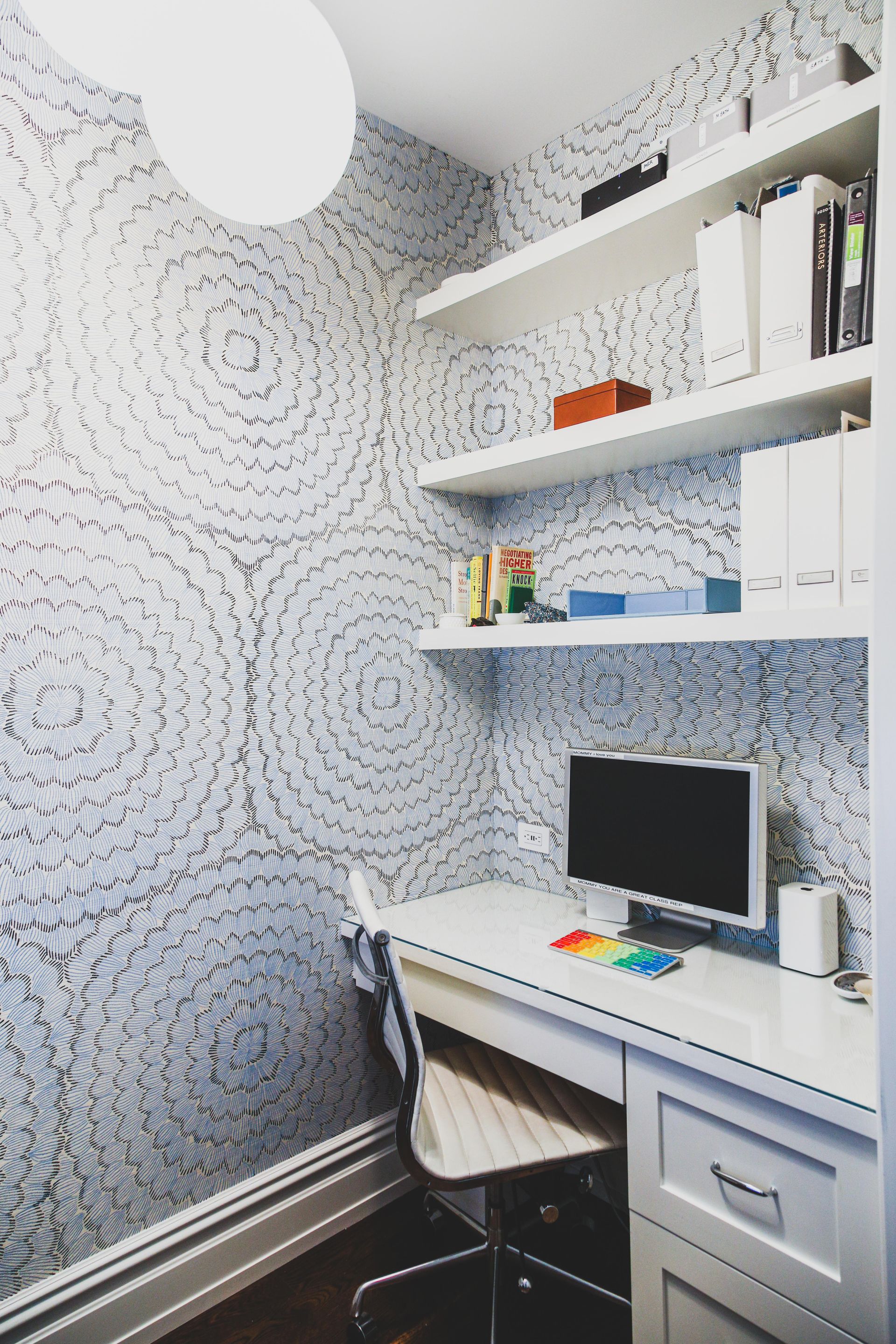 Home office with patterned wallpaper, white desk, shelves, and a computer.