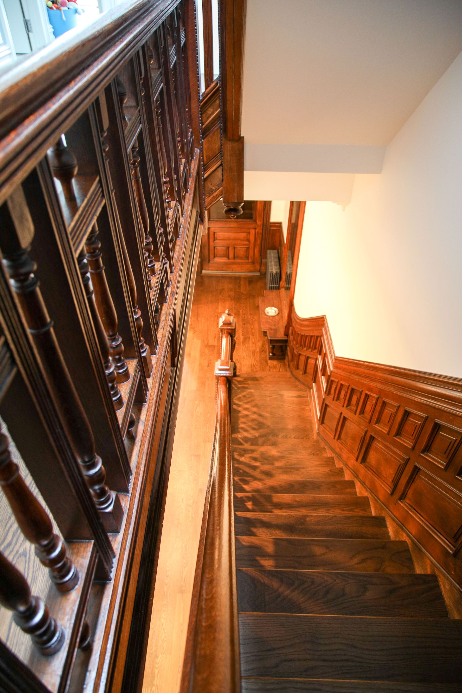 Wooden staircase with ornate banister, viewed from above, descending into a hallway with a doorway.