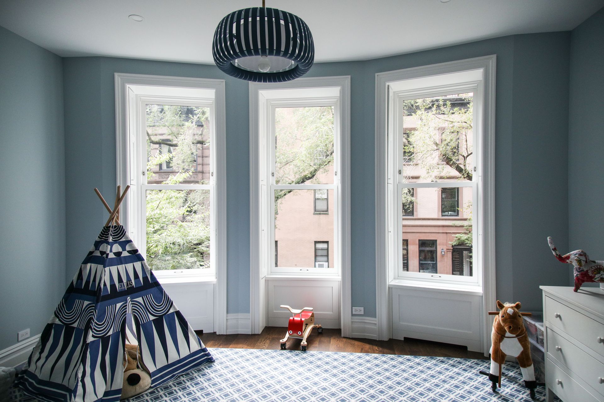 Blue nursery with teepee, rug, and windows overlooking buildings.