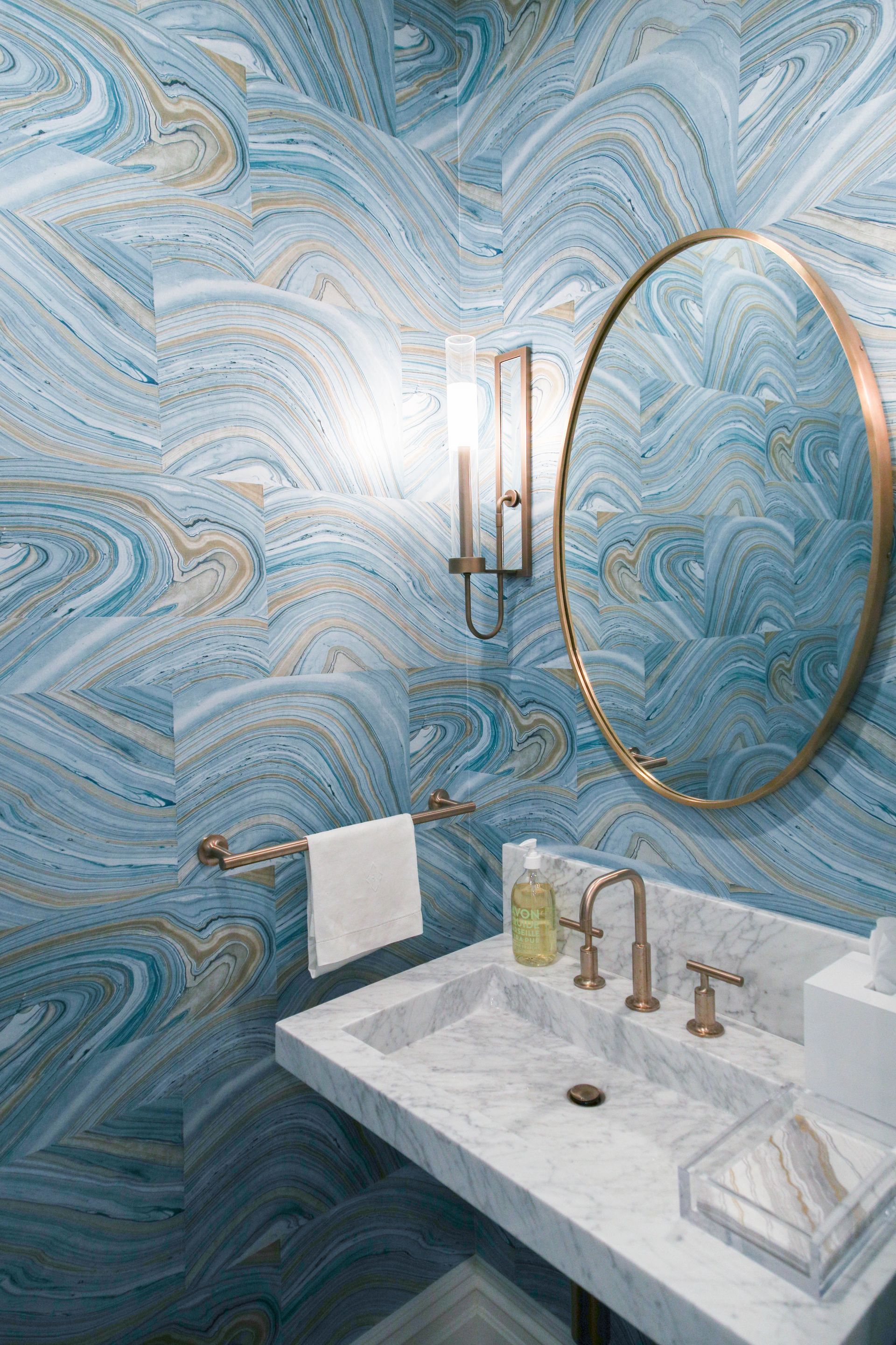Powder room with blue marbled walls, marble sink, gold fixtures, and round mirror.
