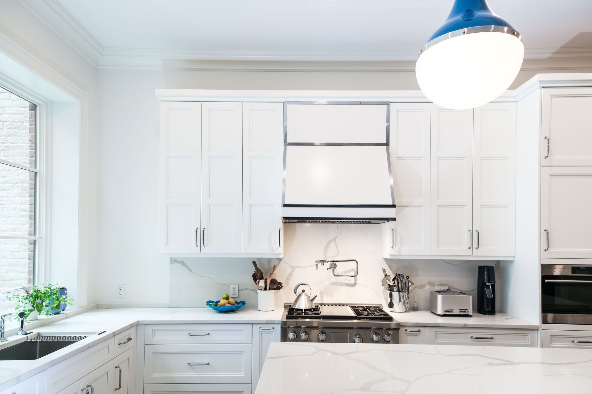 Bright white kitchen with a blue pendant light, marble countertops, and a large window.