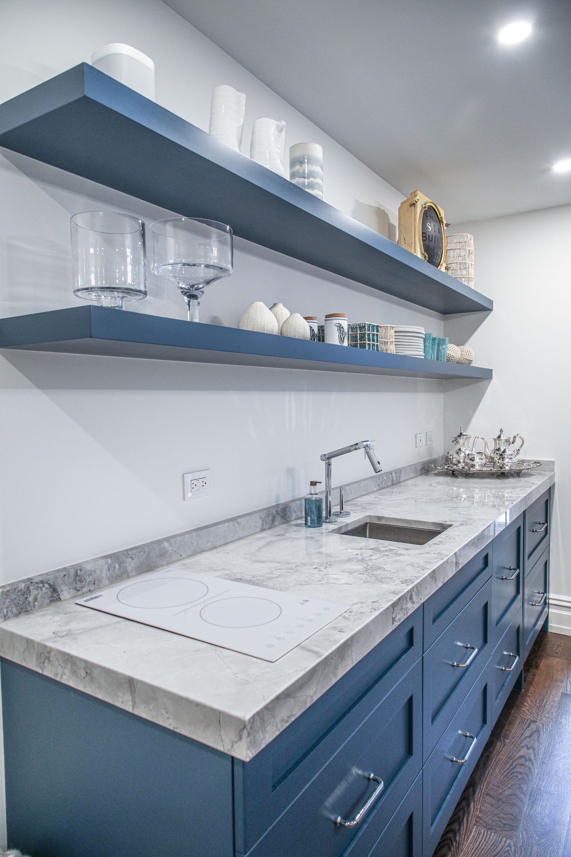 Blue cabinets with marble countertop, sink, and floating shelves against a white wall.