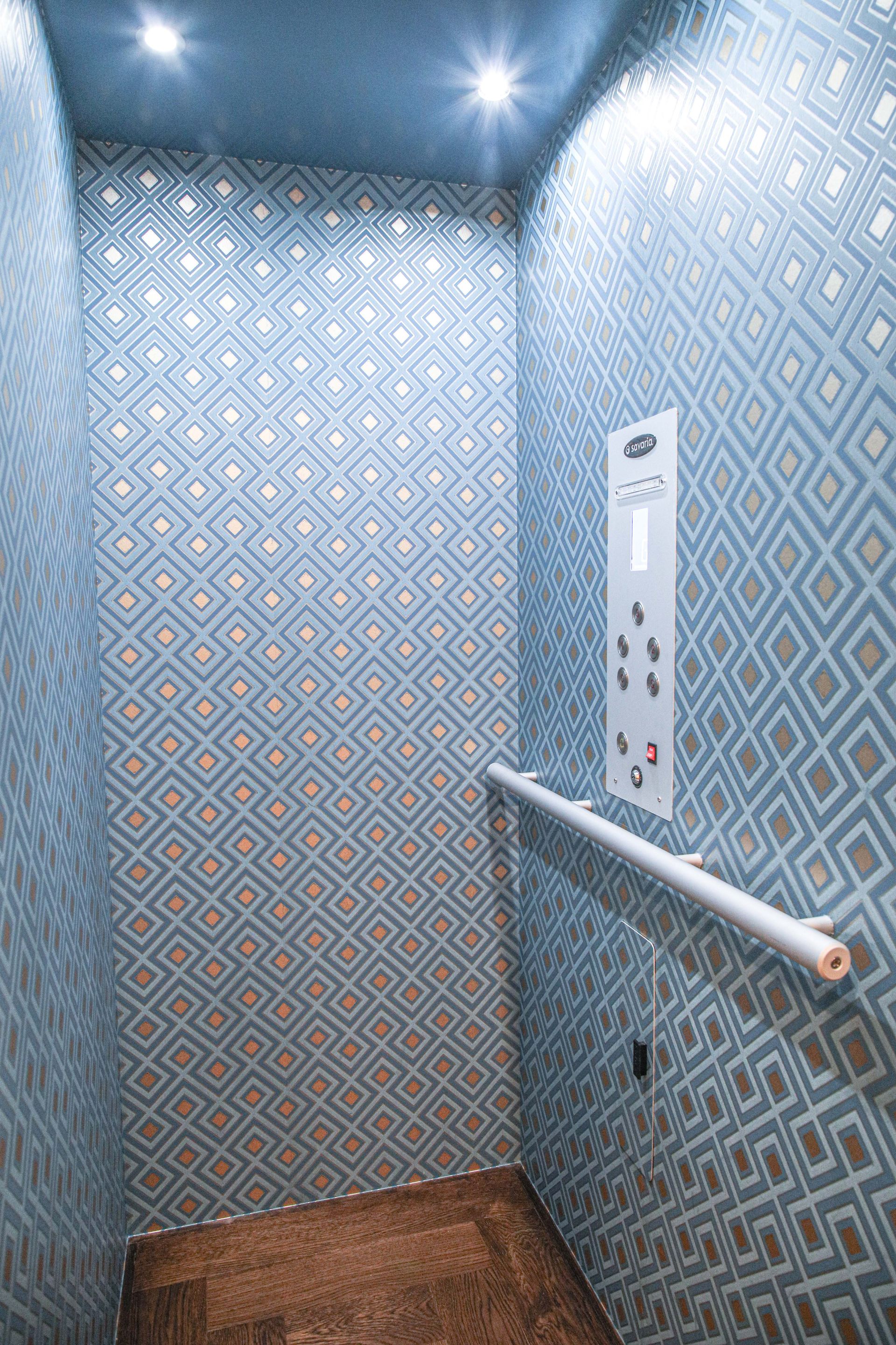 Elevator interior with patterned blue wallpaper, silver accents, and a handrail.