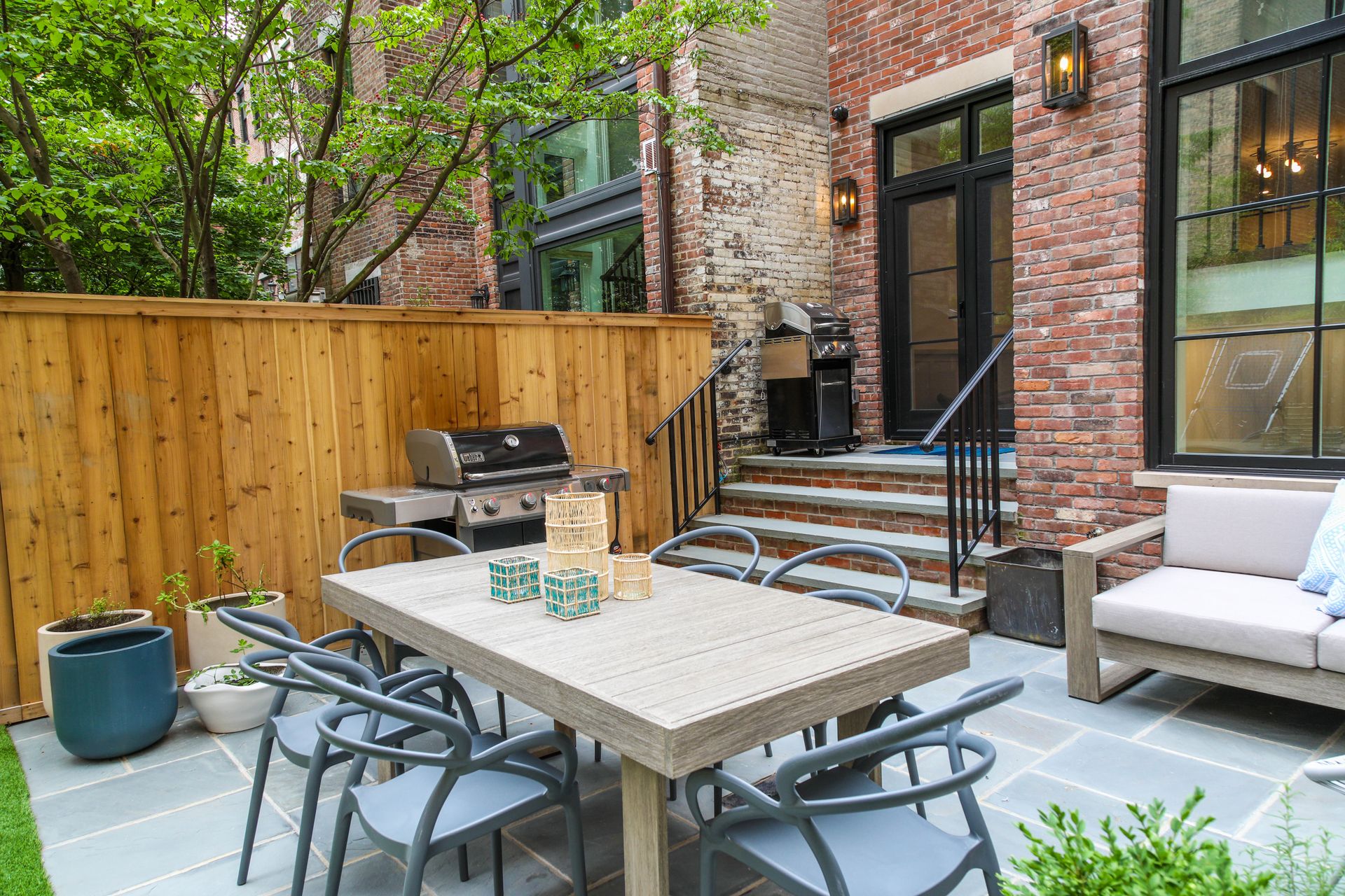 Patio with dining table, grill, and seating. Red brick building and wooden fence surround the space.