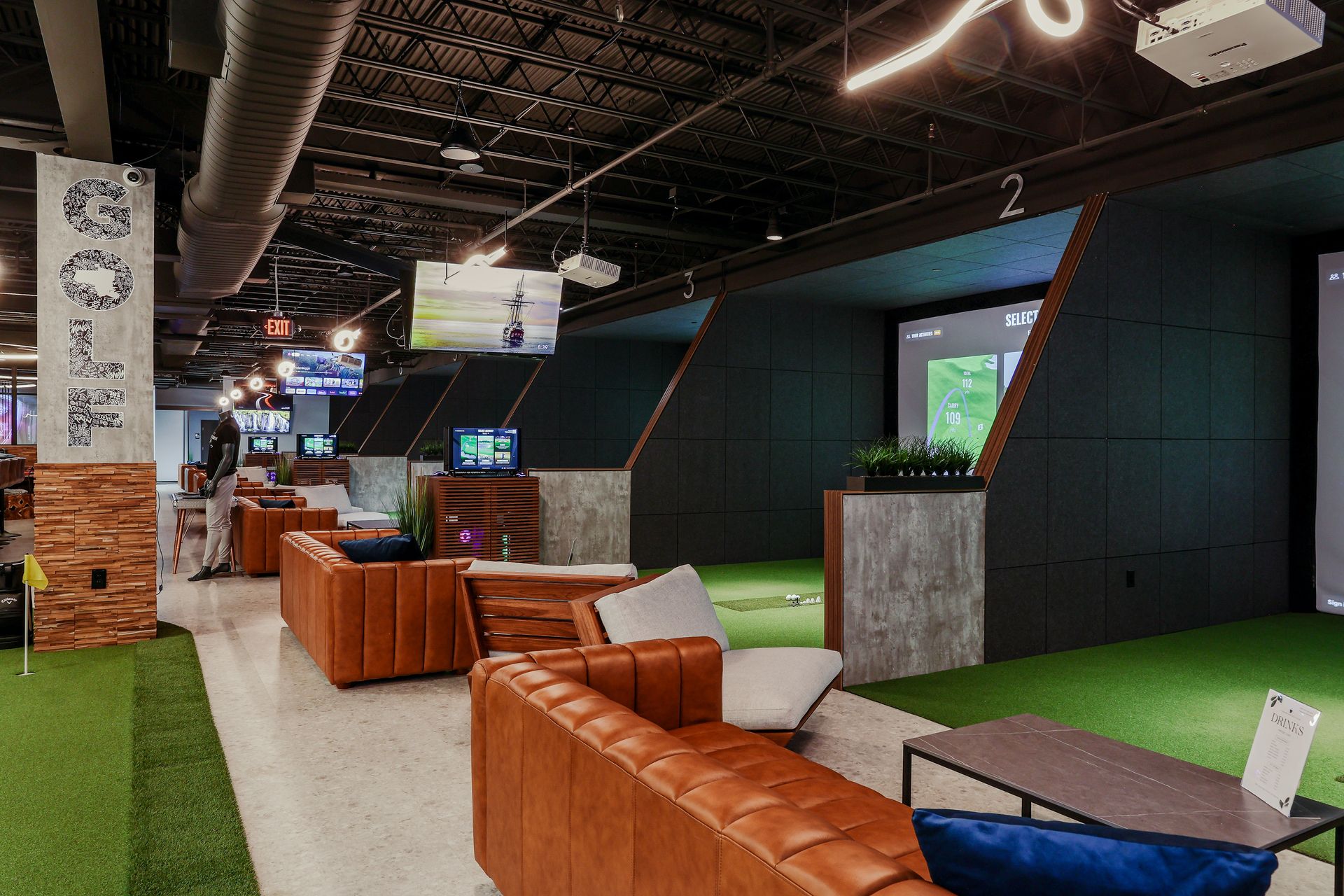 Indoor golf simulator with brown leather couches, green turf, and screens.
