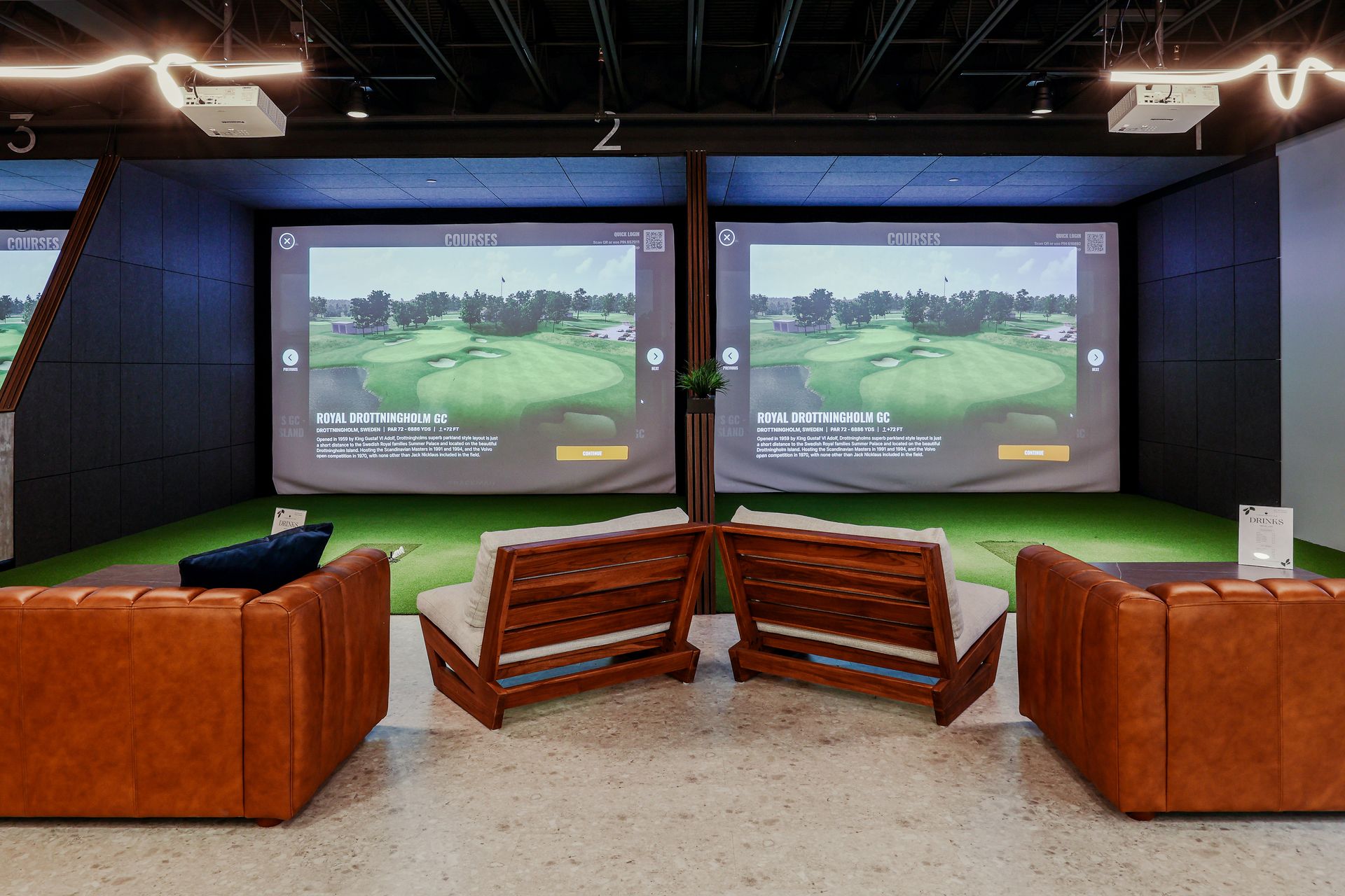 Golf simulator bays with screens, seating, and artificial turf. Two bays are side-by-side, each with a screen displaying a golf course.