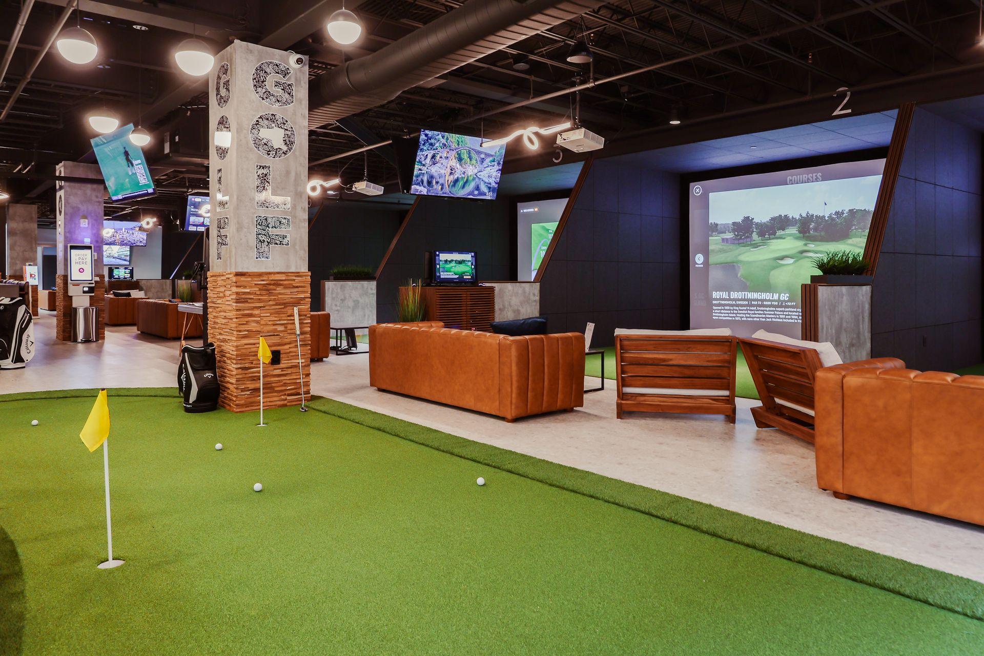 Indoor golf facility with putting green and golf simulator bays, featuring couches and TVs.