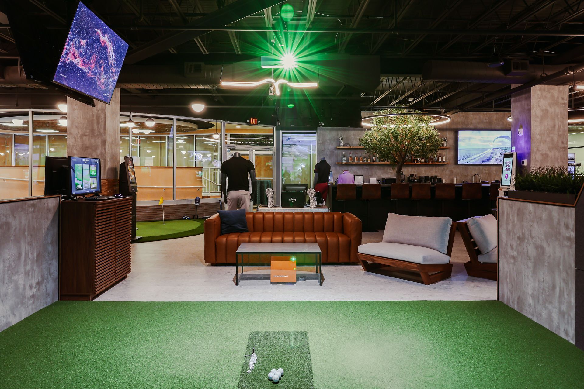 Interior of a golf simulator lounge with turf, seating, screens, and golf balls.