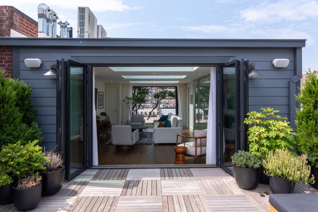Rooftop terrace with open folding doors leading into a bright living room; various potted plants, wood flooring.