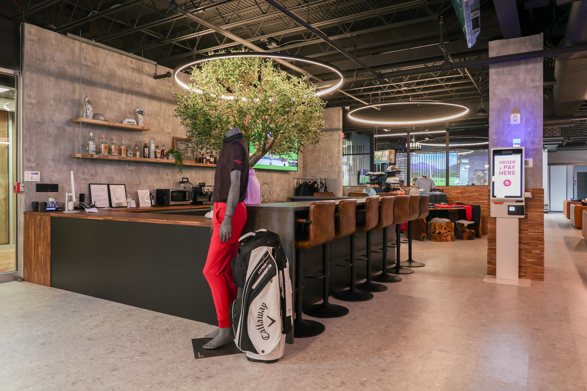 Interior of golf simulator lounge with bar, mannequin in red pants, and golf bag.