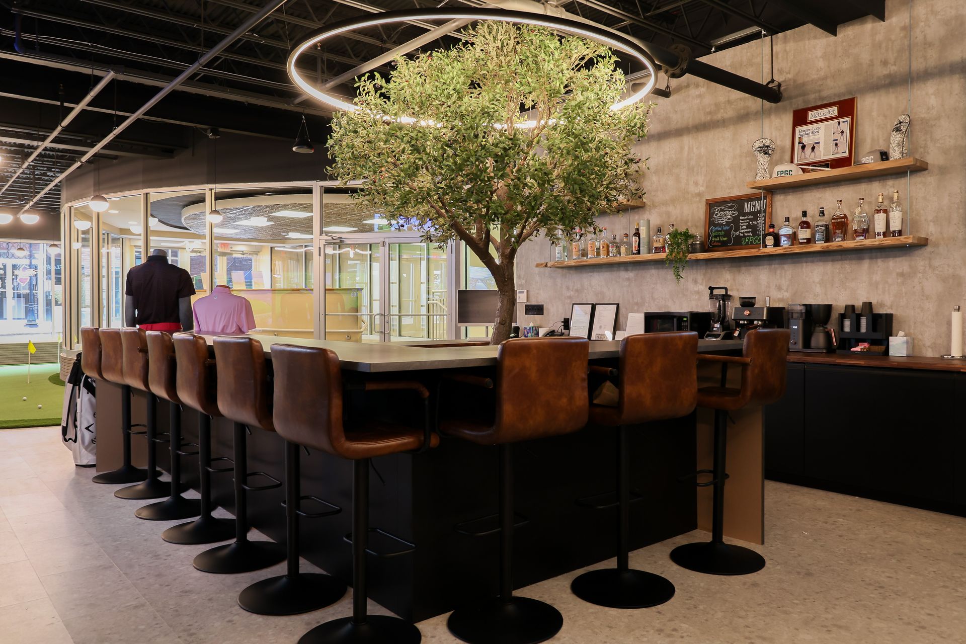 Bar with brown leather stools, an indoor tree, and shelves with bottles, likely in a golf simulator space.