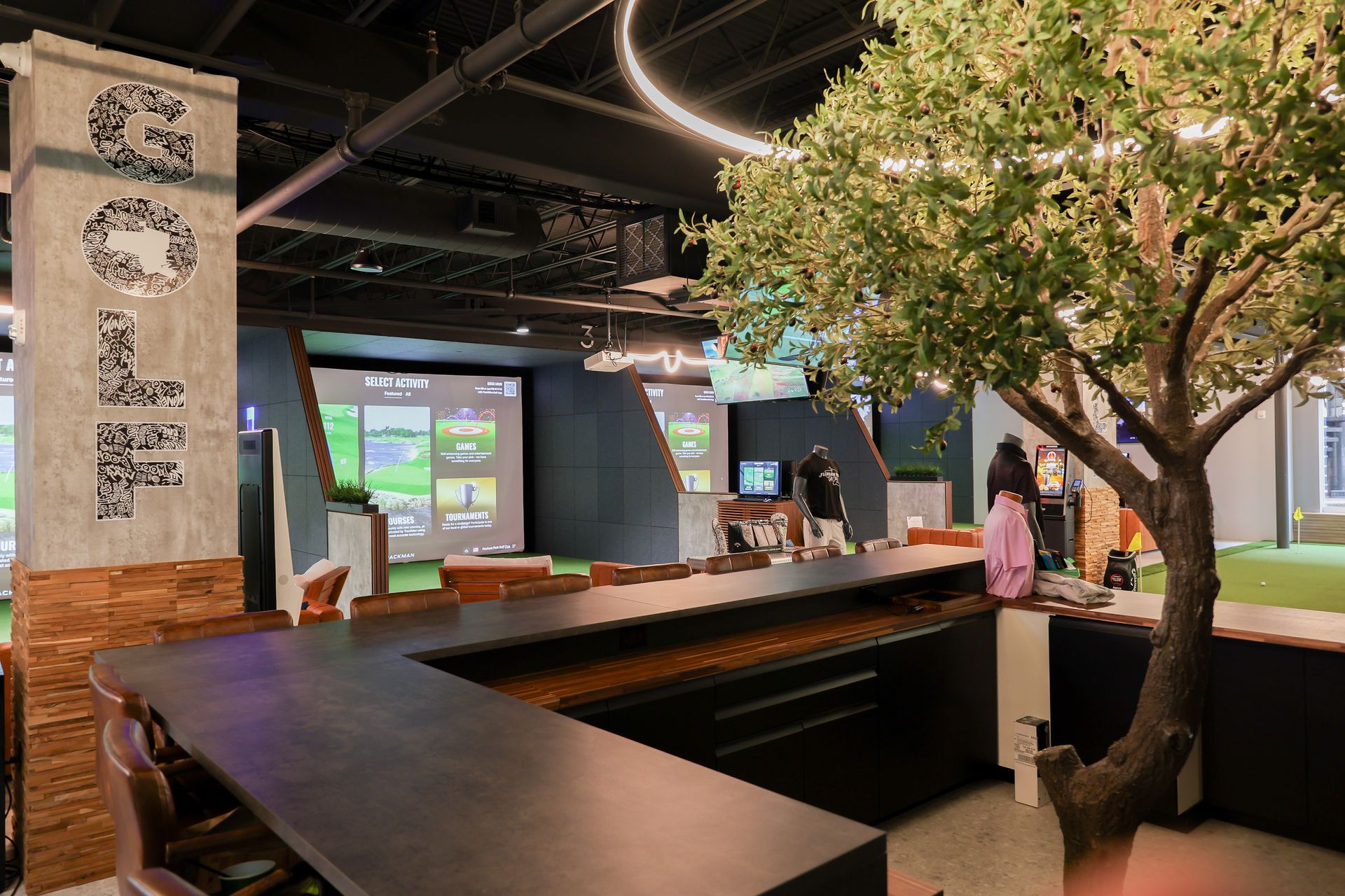 Interior of a golf simulator, featuring screens displaying a course, a bar, and an artificial tree.