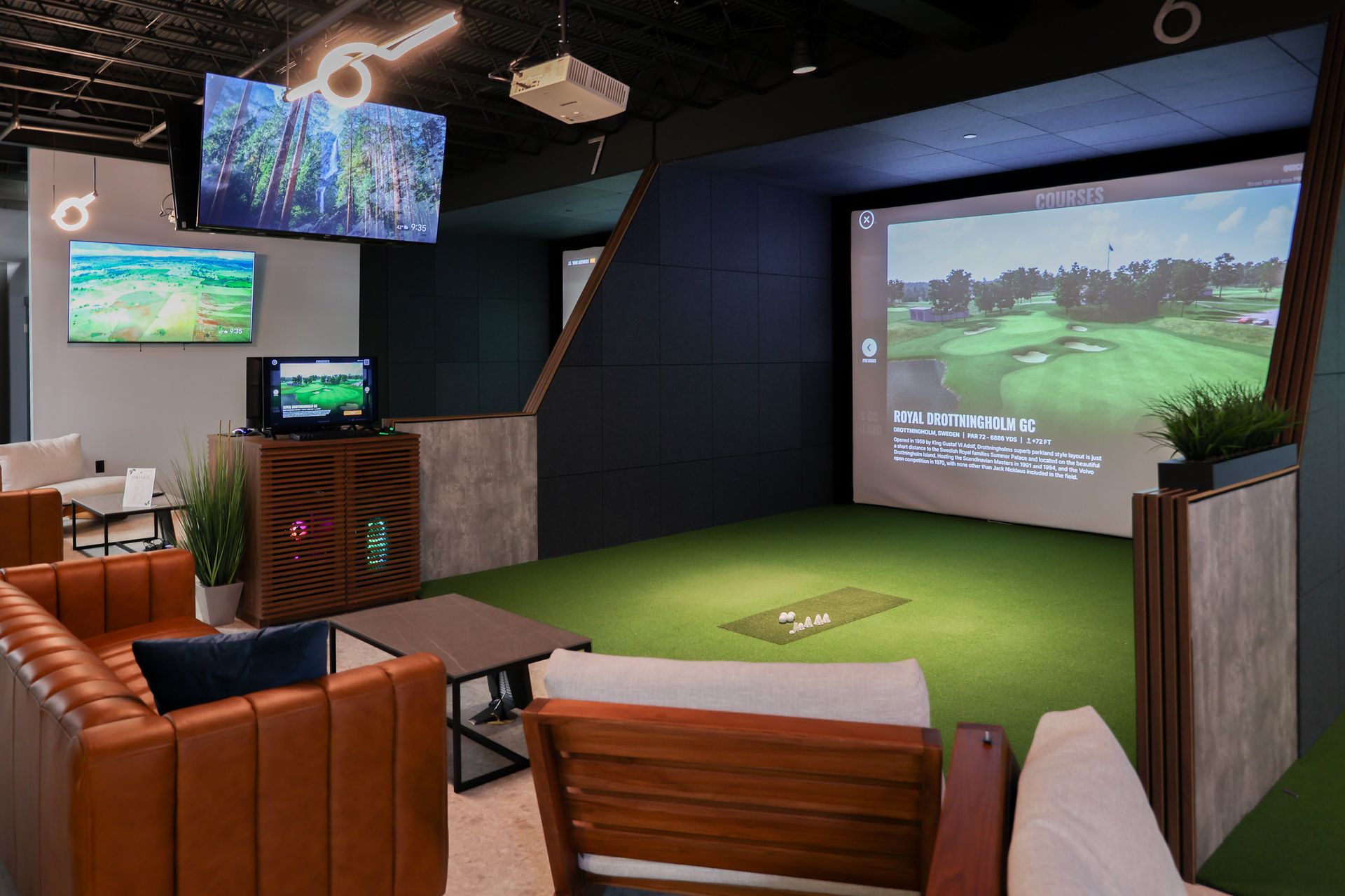 Indoor golf simulator room with golf course image on screen, seating, and multiple TVs.