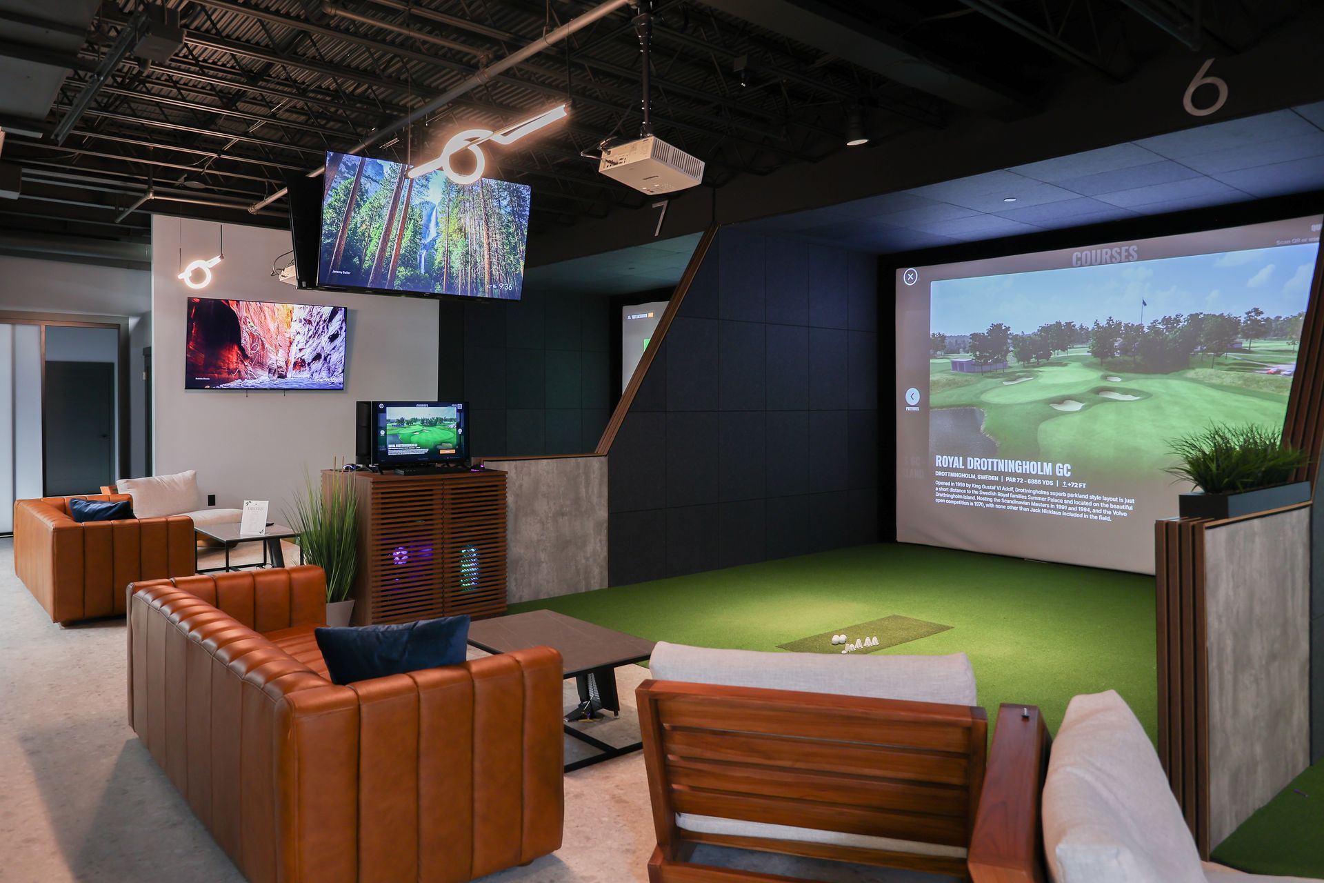 Golf simulator room with screen, artificial turf, couches, and TVs.