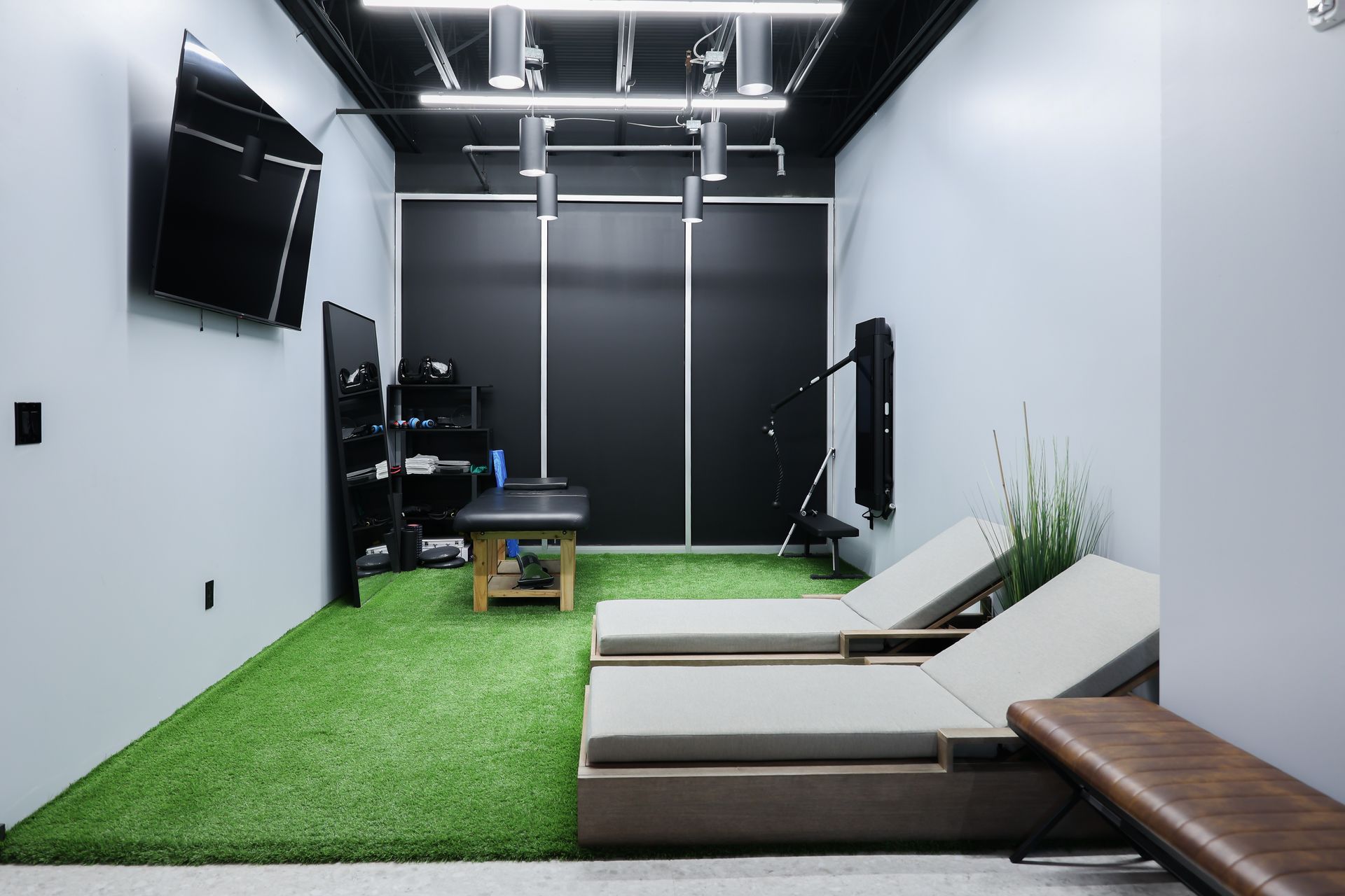 Small gym with turf flooring, two lounge chairs, exercise equipment, and a wall-mounted TV.