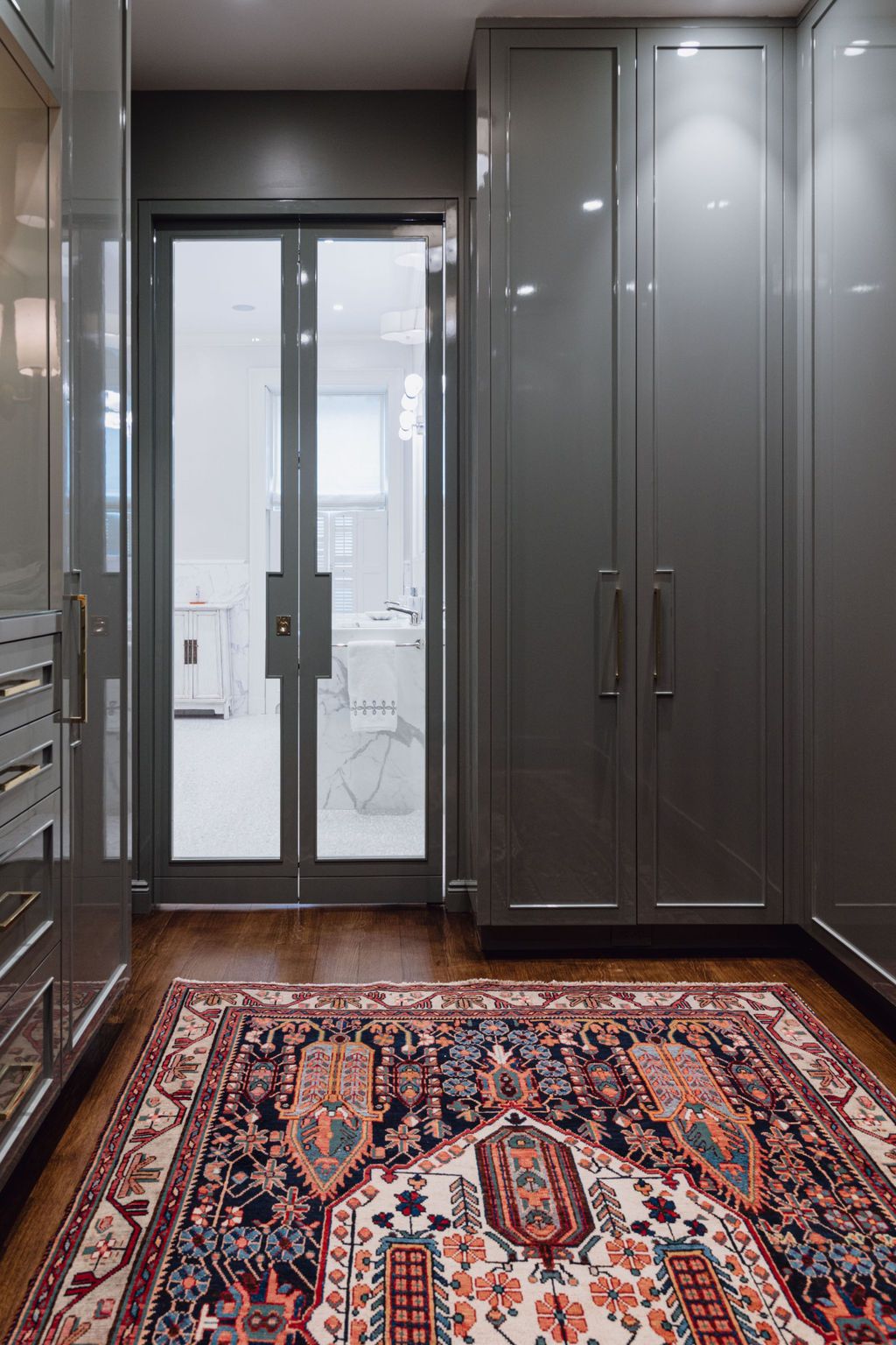 Walk-in closet with dark gray cabinets, oriental rug, and glass doors leading to a white bathroom.