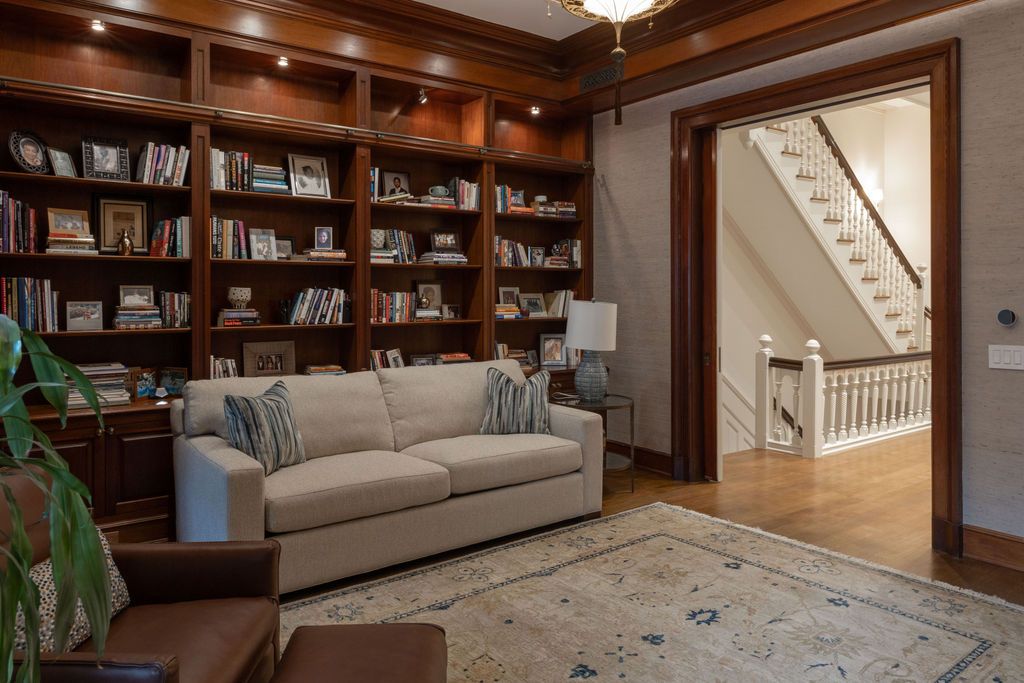 Library with built-in bookshelves, sofa, area rug, and doorway to a staircase. Wooden accents and warm lighting.