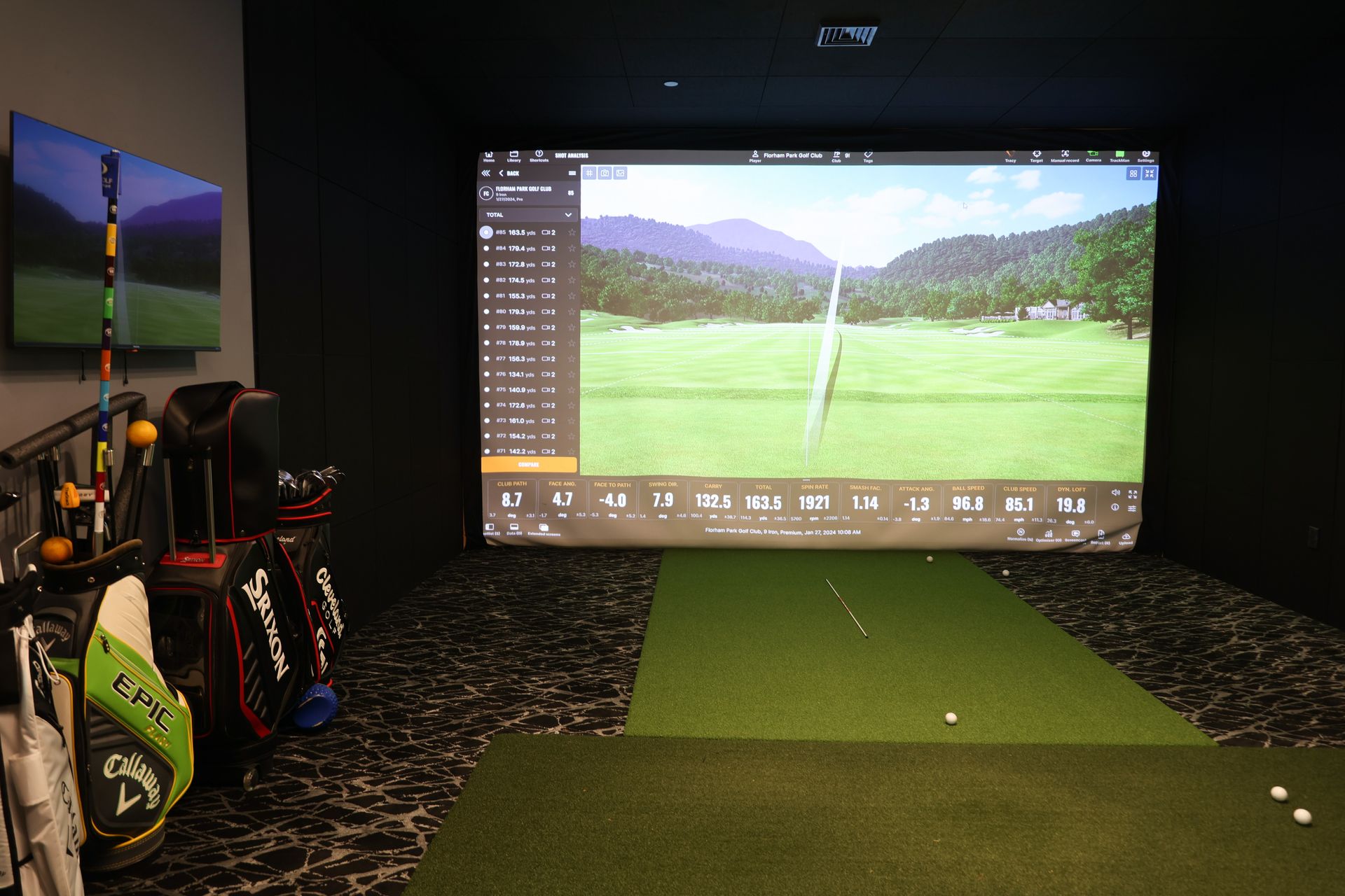 Golf simulator room with screen displaying golf course, golf clubs, and hitting mat.