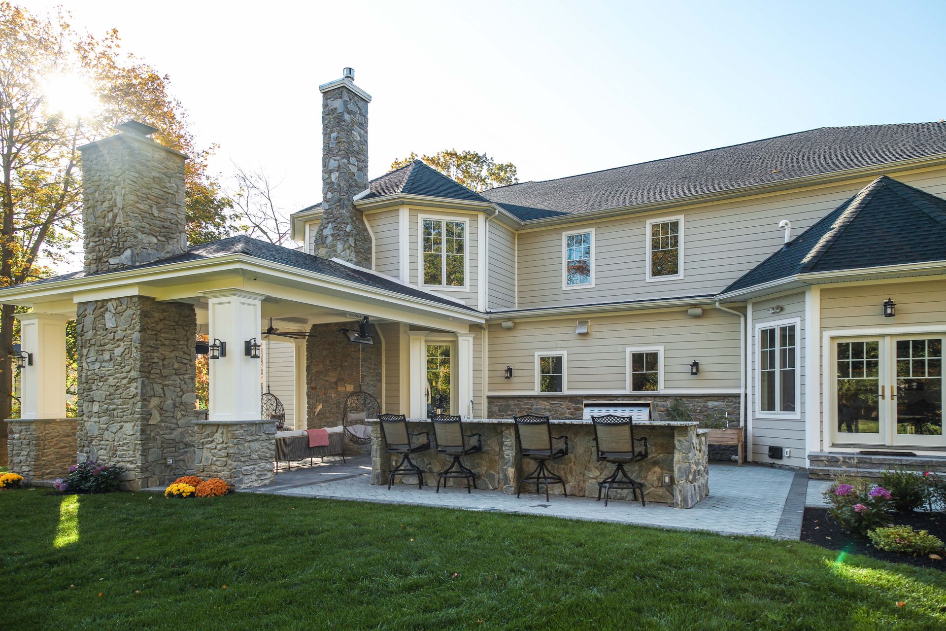Backyard patio with stone bar, seating, and a covered porch extending from a two-story beige house under a sunny sky.