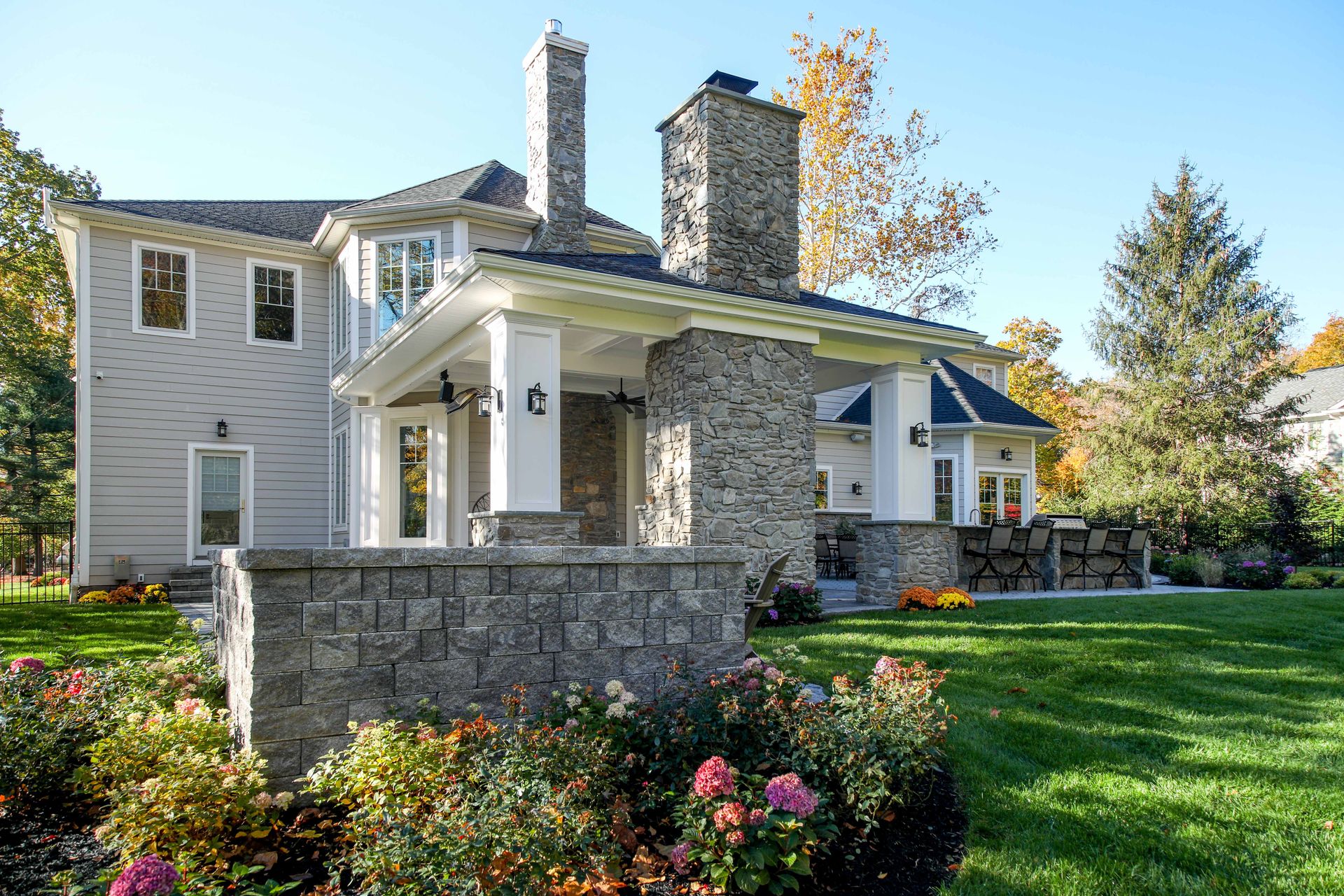 House with stone columns, covered porch, and chimney against a blue sky, surrounded by green lawn and flowers.