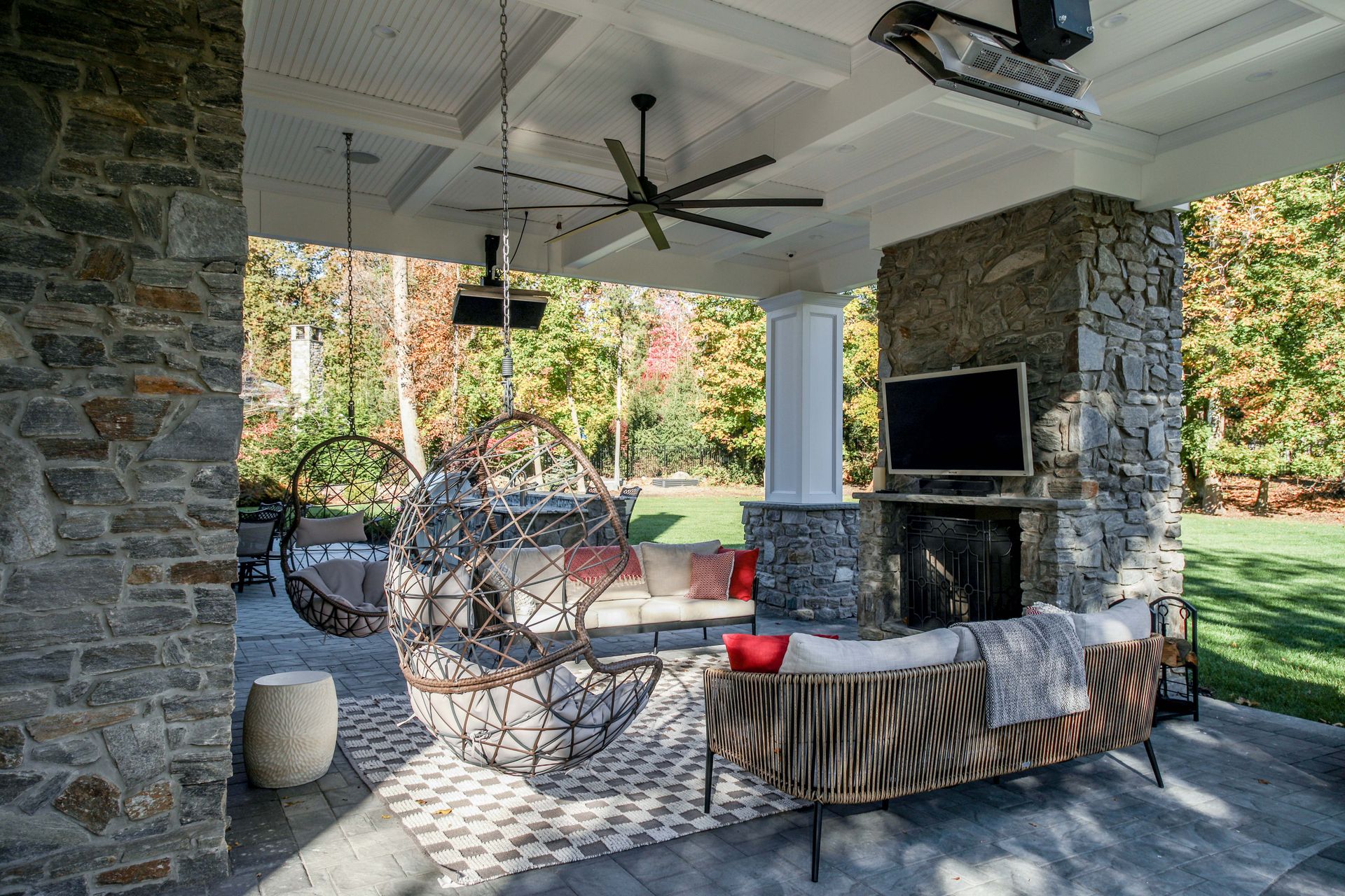 Covered outdoor patio with stone fireplace, seating, hanging chairs, and ceiling fan.