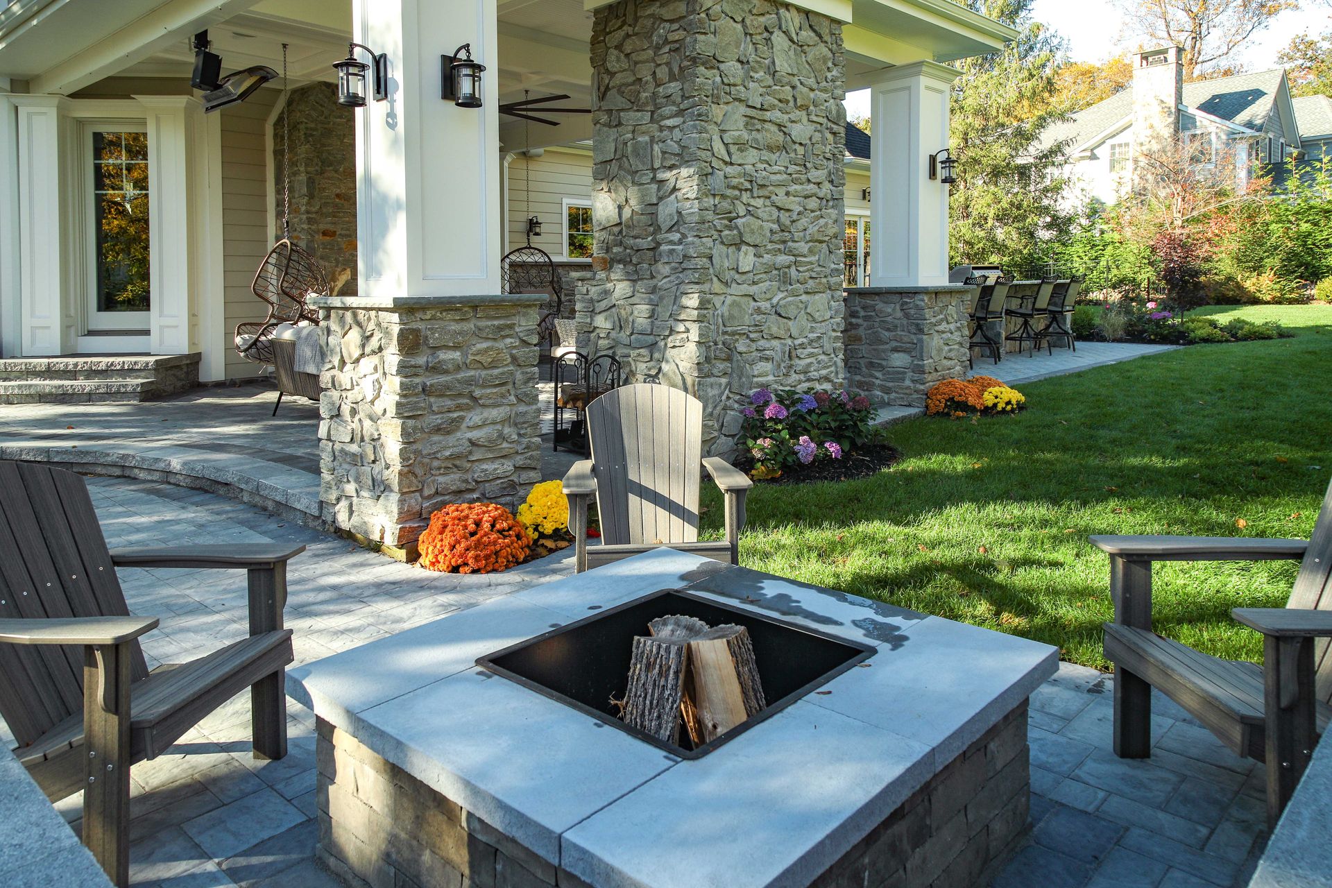 Outdoor seating area with fire pit, stone columns, wooden chairs, and green lawn.