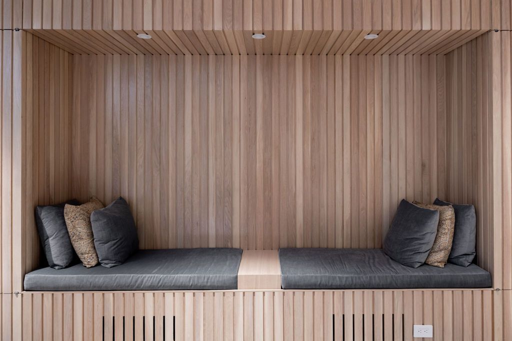 Wooden built-in bench with gray cushions and pillows, recessed in a wooden wall, illuminated by overhead lights.