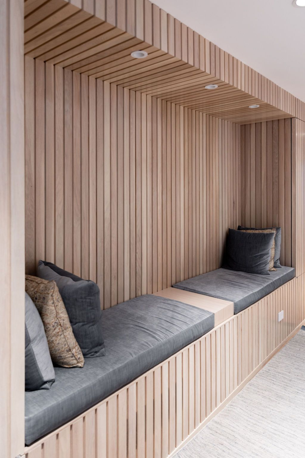 Built-in wooden reading nook with gray cushions and pillows. Recessed lighting.