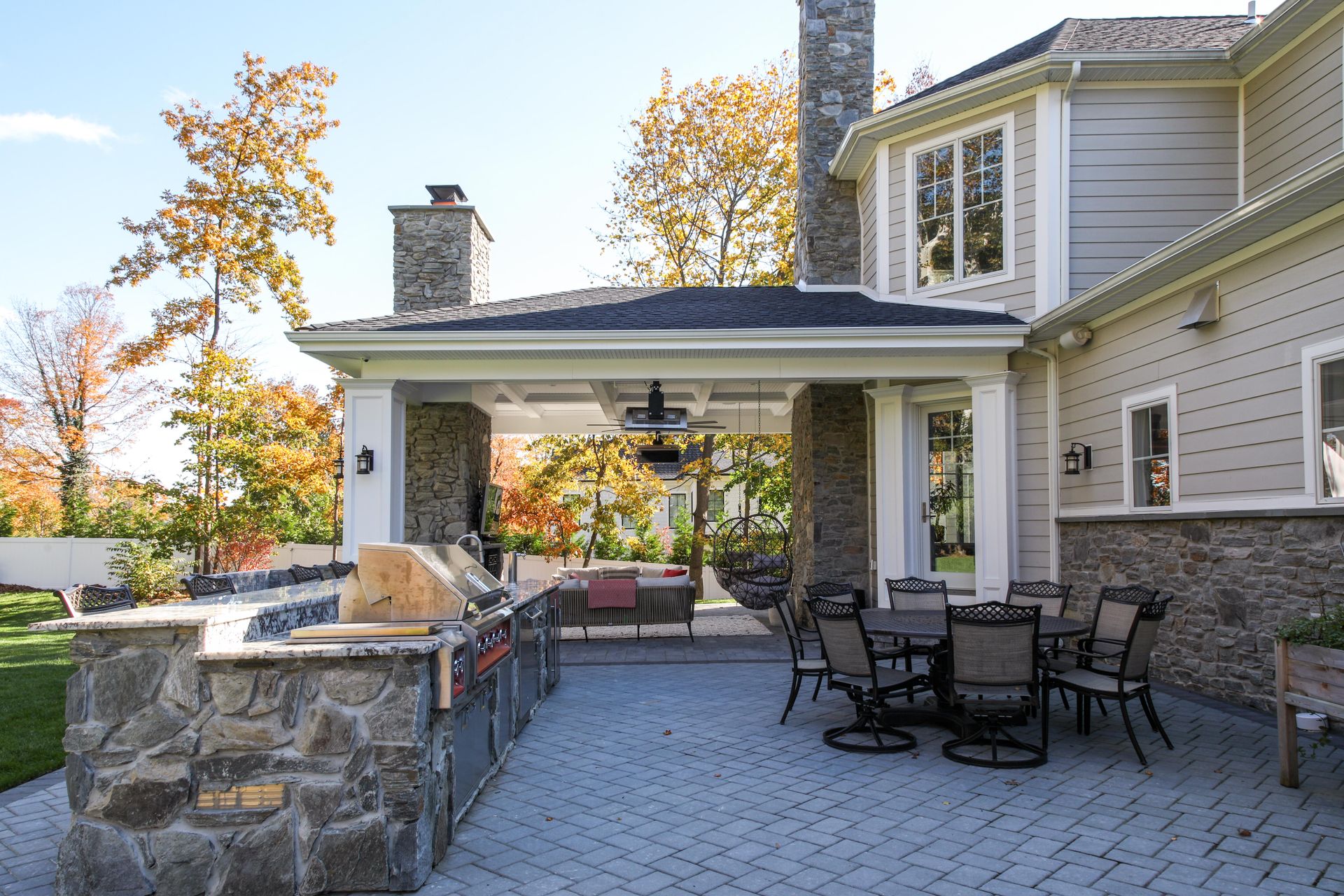 Patio with outdoor kitchen, dining table, and covered seating area, fall foliage in the background.