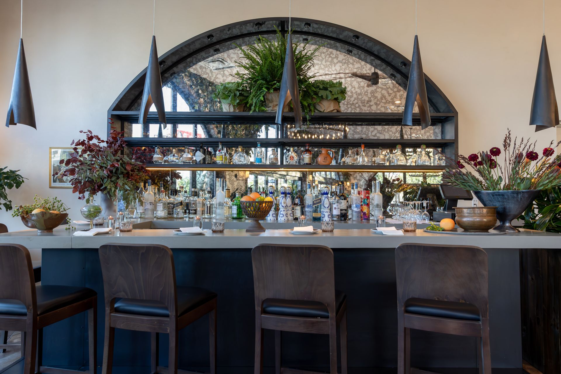 Bar with gray counter, blue base, and brown barstools. Large mirror and plants behind the bar.